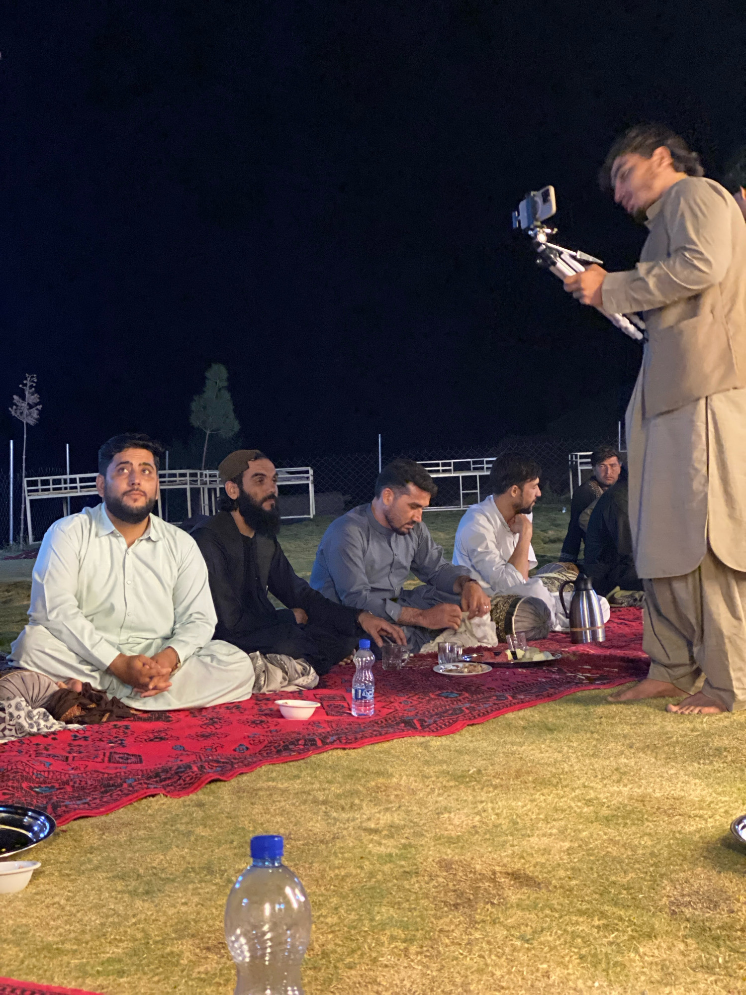 In the city of Gul Kalachah, Afghanistan, a group of men are gathered outdoors at night on a red and black patterned rug laid on a grassy area. The men are seated, facing towards the left of the frame. In the foreground, a man wearing a light blue shalwar kameez is seated with his hands clasped in his lap. To his right, a man in a dark shalwar kameez with a brown cap is looking to his right. Further to the right, a man in a white shalwar kameez is looking away from the camera, resting his chin on his hand. Another man in a grey shalwar kameez is seated closer to the right edge of the rug, engaged in an activity with his hands. A silver thermos and a plate with food are visible on the rug. A few glasses and a bottle of water are also present. In the background, a man is standing and holding a smartphone on a tripod, appearing to film. He is wearing a light brown shalwar kameez. The environment appears to be a courtyard or an open area with some fencing and trees visible in the dark night sky.