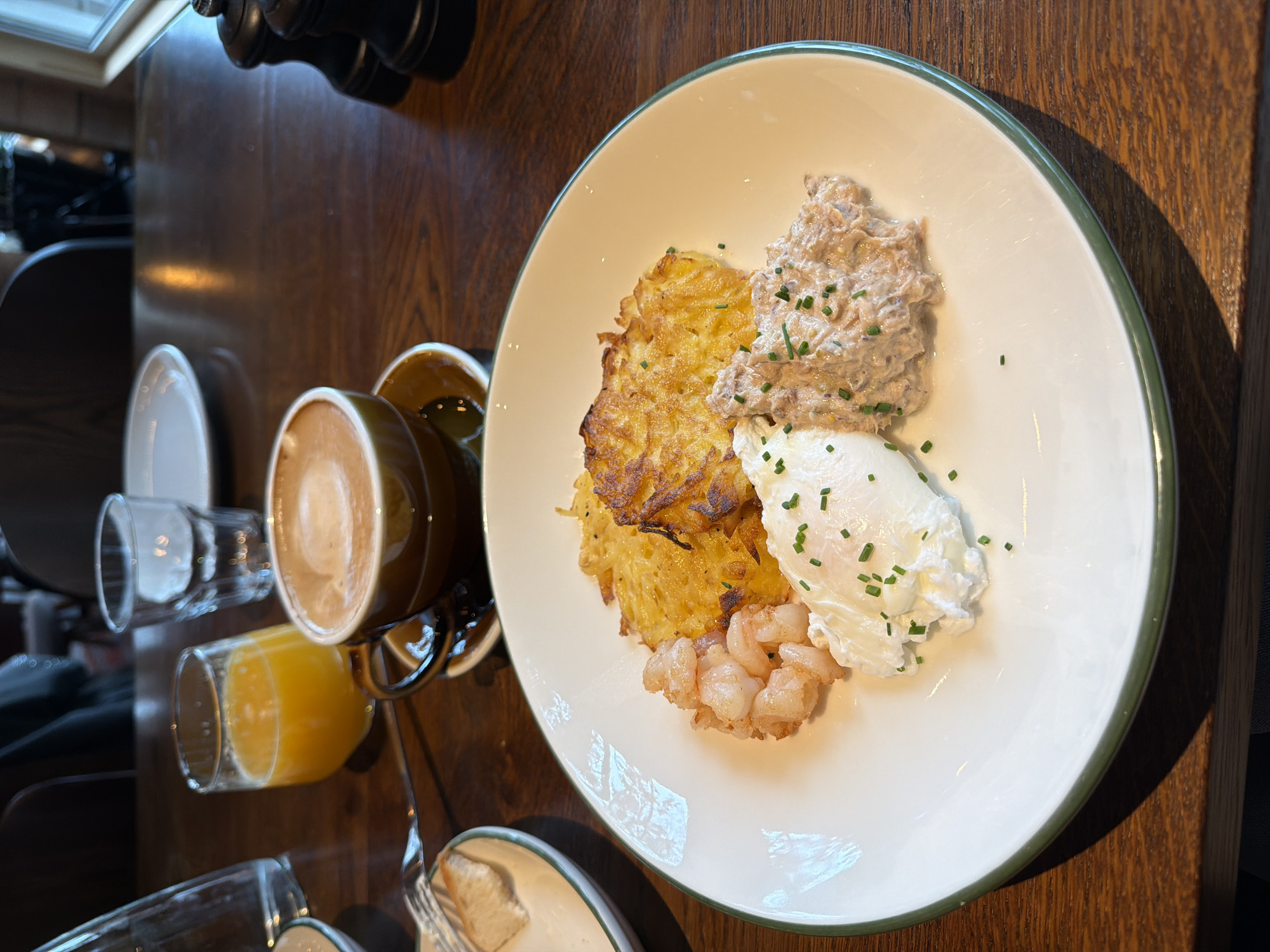 A high-angle shot shows a plate of food on a wooden table alongside beverages. The plate contains a potato cake, a dollop of what appears to be salmon salad, poached eggs sprinkled with chives, and cooked shrimp. Next to the plate is a cup of cappuccino and a glass of orange juice, as well as what appears to be a second glass with liquid inside. The location is a restaurant or cafe in Санатория "Восход", Russia.