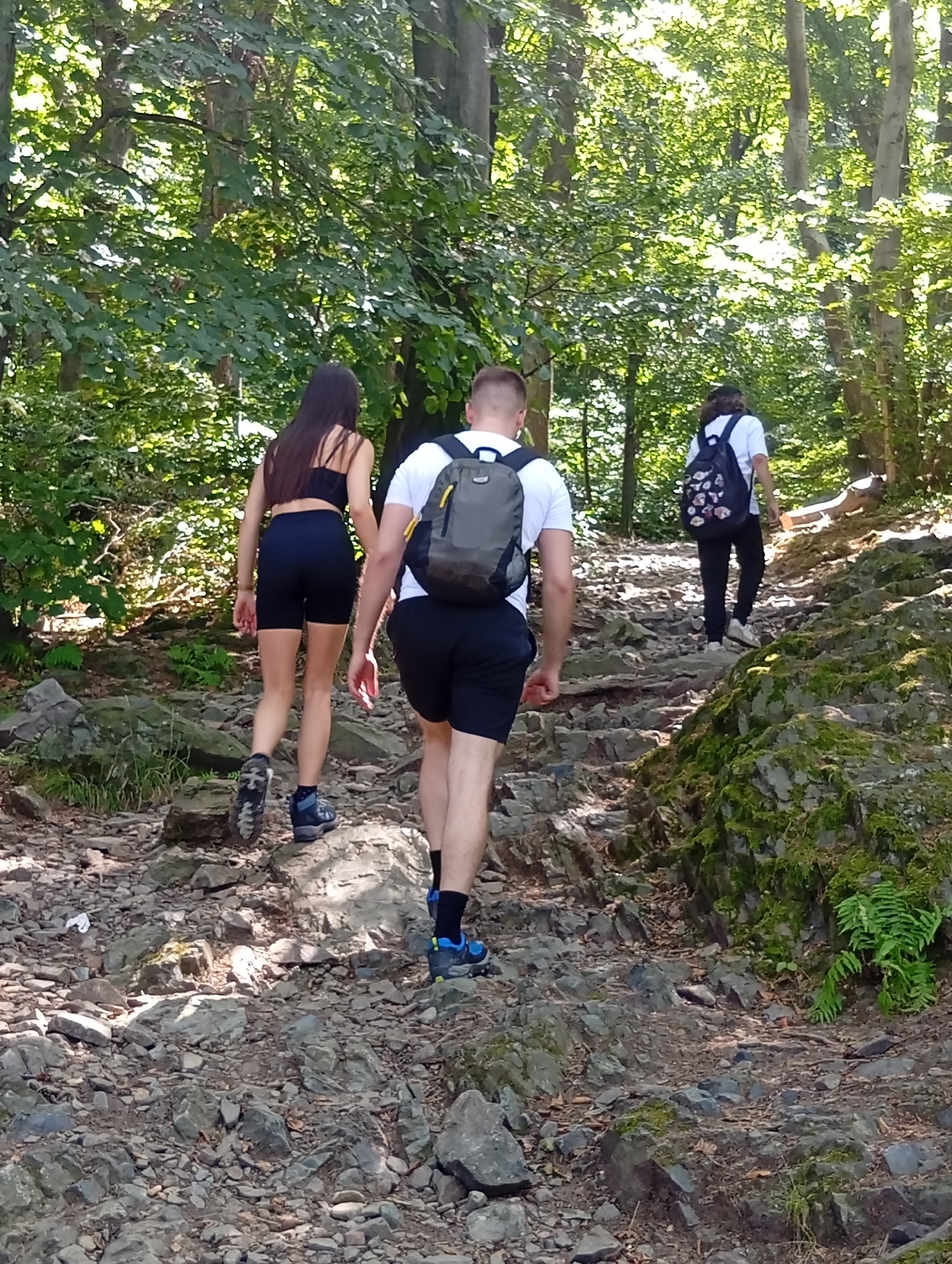 Three individuals are hiking up a rocky, wooded path. The person in the foreground on the right is a male wearing a white t-shirt, black shorts, black socks, and blue hiking shoes with a grey backpack. To his left, a female is wearing a black sports bra, black shorts, and blue hiking shoes, with her back to the camera. Further up the path and to the right, another person, likely female, is wearing a white t-shirt, black pants, white sneakers, and a backpack adorned with floral patterns. The environment is a forest with trees and dappled sunlight, suggesting a temperate climate. The path is uneven and covered in stones and moss. The scene appears to be on Mount Ślęża near Sobótka, Poland, a popular hiking destination known for its natural beauty and rocky terrain.