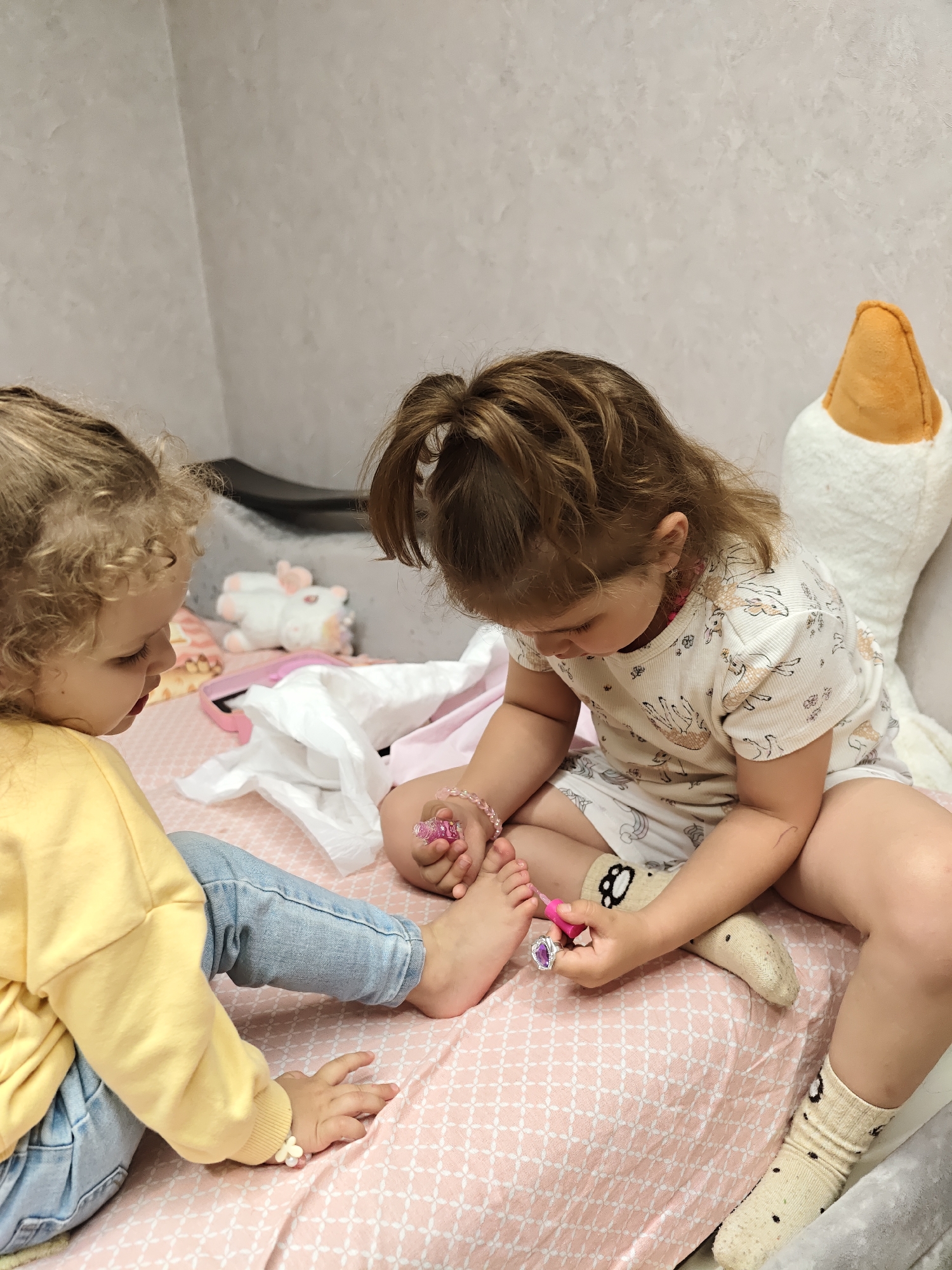 Two young children are on a bed with a pink patterned blanket. The child on the left has curly blond hair, wears a yellow sweater and blue jeans, and appears to be watching. The child on the right has brown hair in a ponytail, wears a white patterned short-sleeved pajama set and socks with cartoon faces on them, and is applying pink nail polish to the toes of the first child. A plush goose and other toys are in the background. The setting appears to be an indoor room with light-colored walls. There is no information about the specific location in the city of Chaikovsky, Russia.
