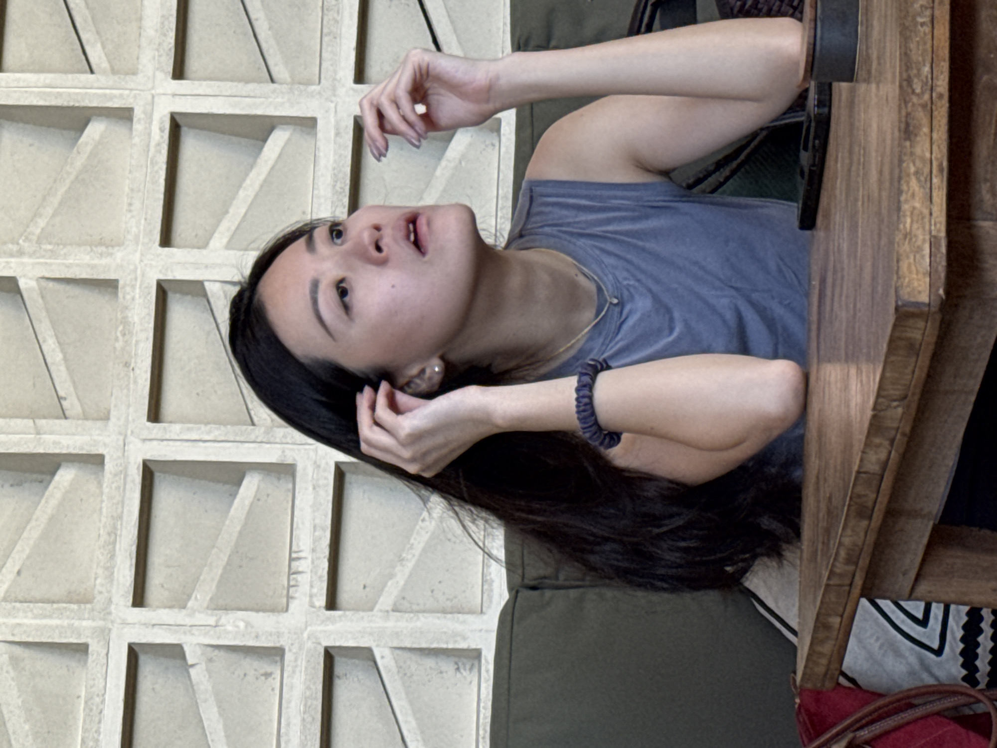 A woman with long dark hair, wearing a blue t-shirt and a beaded bracelet, is seated at a wooden table. She is looking upwards with her mouth slightly open and her hands near her face. Behind her is a wall constructed of textured rectangular blocks with a geometric design, and below her is a green cushion. The setting appears to be indoors, possibly a cafe or restaurant. The exact location within Kuta Selatan, Indonesia is not determinable from the image.