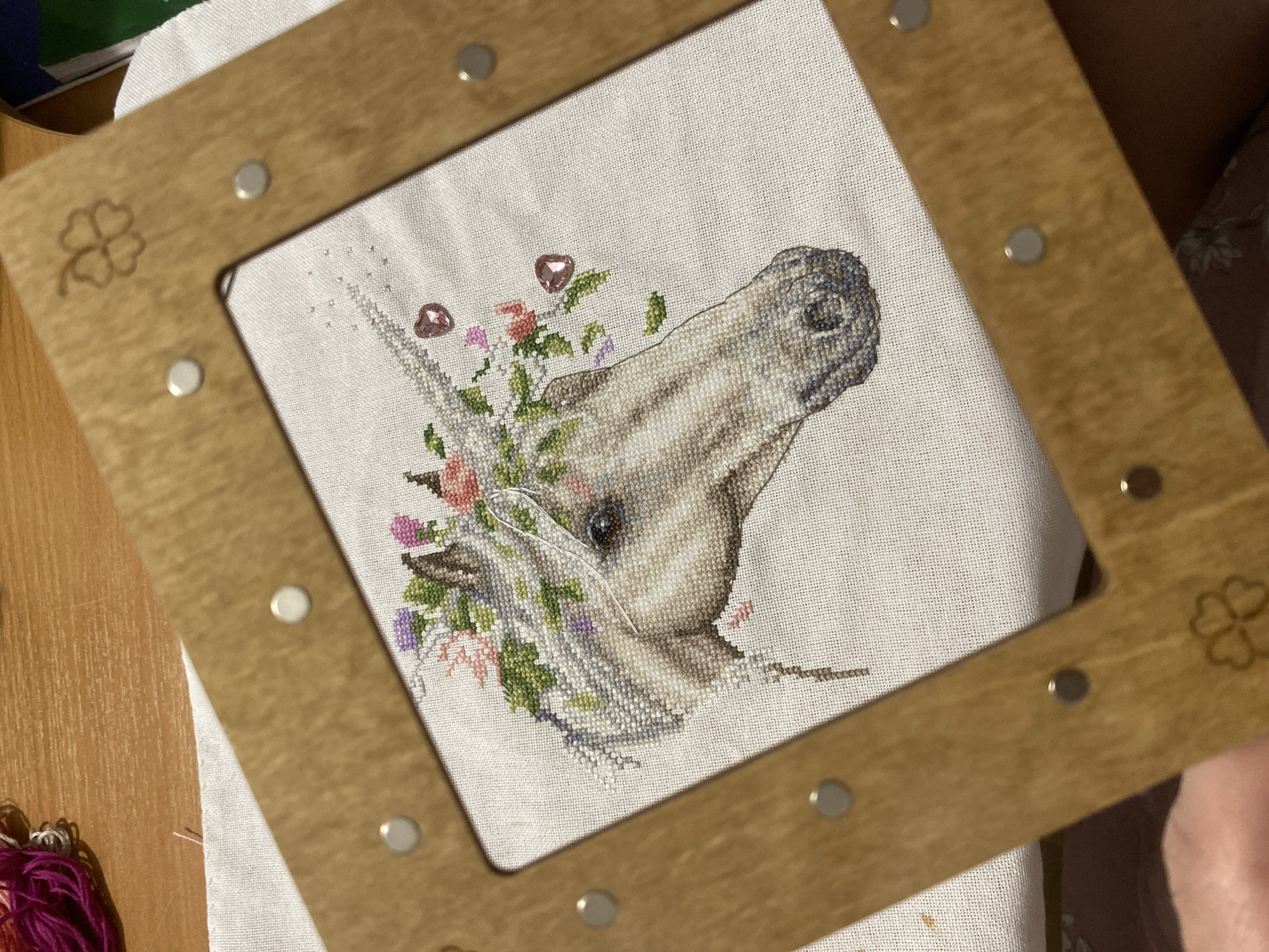 A close-up, slightly angled shot depicts a cross-stitch artwork of a horse head adorned with flowers, held within a wooden frame. The frame has decorative cloverleaf designs in two corners and metallic studs along its edges. The cross-stitch work shows the horse in profile, facing right, with details including shading to define its form and texture. Flowers are stitched around its head and neck. Two shiny, heart-shaped embellishments are affixed to the embroidery, possibly representing jewels or decorative accents. A needle and thread are partially visible within the stitched artwork. The fabric onto which the cross-stitch is made is a light color, likely white or off-white. The wooden frame appears to be a light brown color. Part of a table is visible below left and to the right is a portion of clothing on a person's lap. The location of Yekaterinburg, Russia is not apparent from the image.