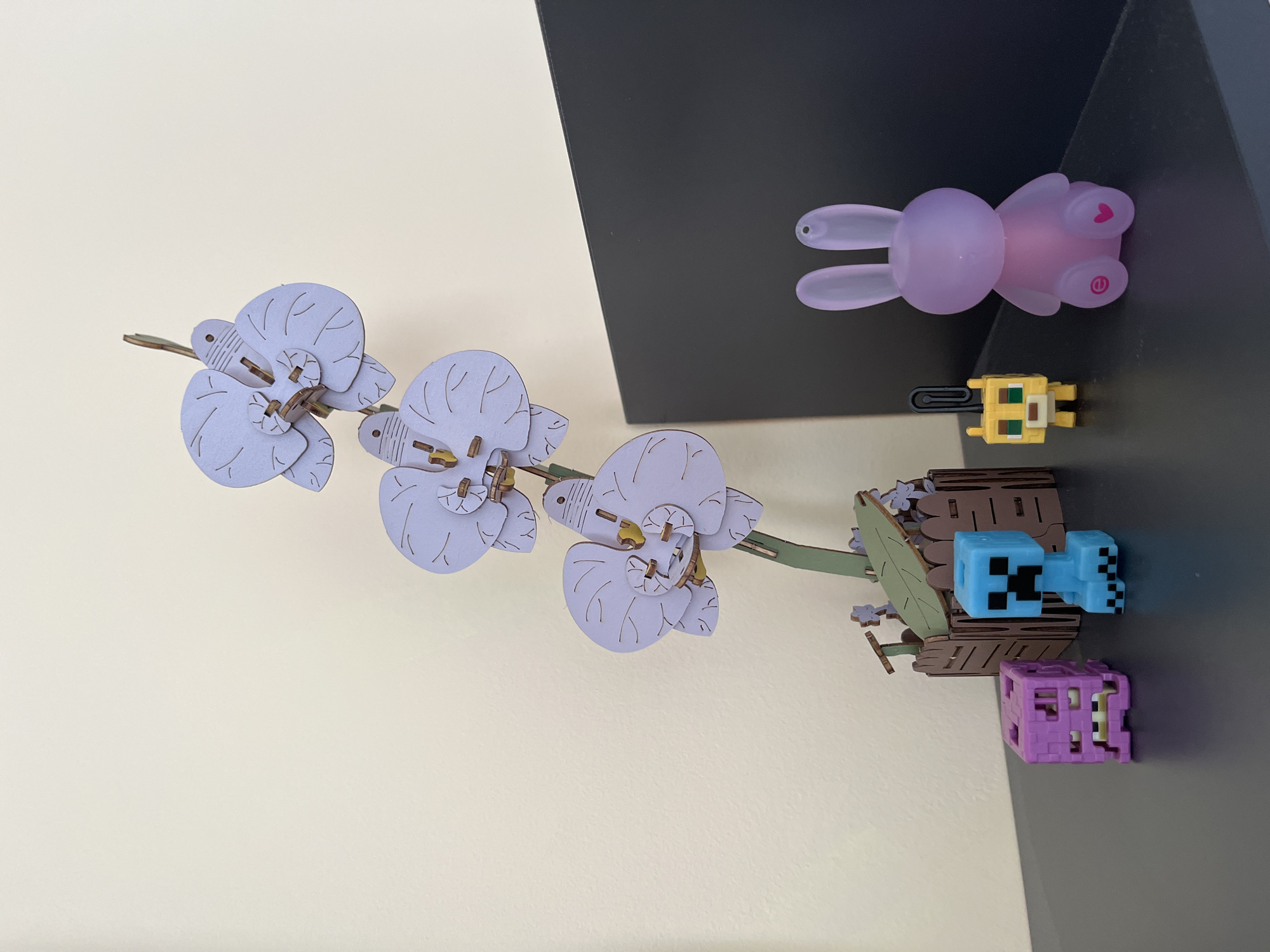 This image displays a collection of small, colorful objects arranged on a wall with a cream-colored section and a dark gray section. The objects appear to be decorative pieces, possibly from a puzzle or toy set.
On the left side, a cluster of light purple orchid-shaped ornaments is attached to a thin, green stem. These ornaments are constructed from layered wood or cardstock, with delicate lines etched onto their petals to mimic natural veins.
To the right of the orchids, on the dark gray surface, several small figurines are positioned. At the top, a translucent, light purple bunny figurine is lying on its back. Below it, a yellow, pixelated figurine, resembling a character from a video game, is standing. Further down, a light blue, pixelated figurine, also reminiscent of a video game character, is placed within a wooden-like structure that appears to be a small chest or crate. Beneath this structure is a purple, cube-shaped object with visible internal details.
The overall arrangement suggests a playful or whimsical display. The lighting is even, and there are no strong shadows, indicating an indoor setting, likely a room. The orientation of the image is such that the objects appear to be on a vertical surface, with the cream and dark gray sections dividing the background. No specific time of day, weather, or emotions are discernible. There is no visible text in the image. The location context provided is Plovdiv, Bulgaria, but this cannot be visually confirmed from the image itself.