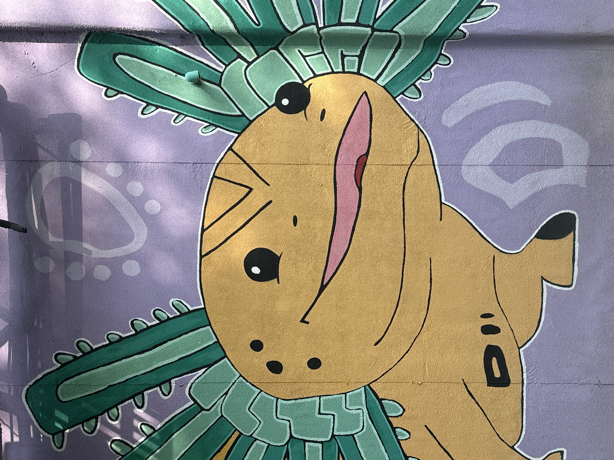A vibrant mural is painted on a textured, light purple wall, featuring a cheerful, cartoonish creature. The central subject is a stylized, yellow-gold character, reminiscent of a fish or an amphibian, with a large, oval head and a smaller body extending to the right. It has prominent black eyes with white pupils, and a wide, open mouth with a pink interior and a small red tongue visible.
The character is adorned with a elaborate green headdress or collar that fans out around its head, composed of multiple spiky or leafy segments in varying shades of green, outlined in black and white. The background of the mural is a soft lavender or light purple, decorated with subtle, light grey geometric and organic patterns, including arcs, circles, and abstract shapes.
The artwork is illuminated by bright daylight, casting distinct shadows from an unseen object, possibly a fence or foliage, across the left side of the wall and parts of the mural itself. The rough texture of the wall is visible through the paint. The character's expression is one of jovial happiness or playfulness. There is no visible text in the image. This piece of public art is located in San Andrés Cholula, Mexico.