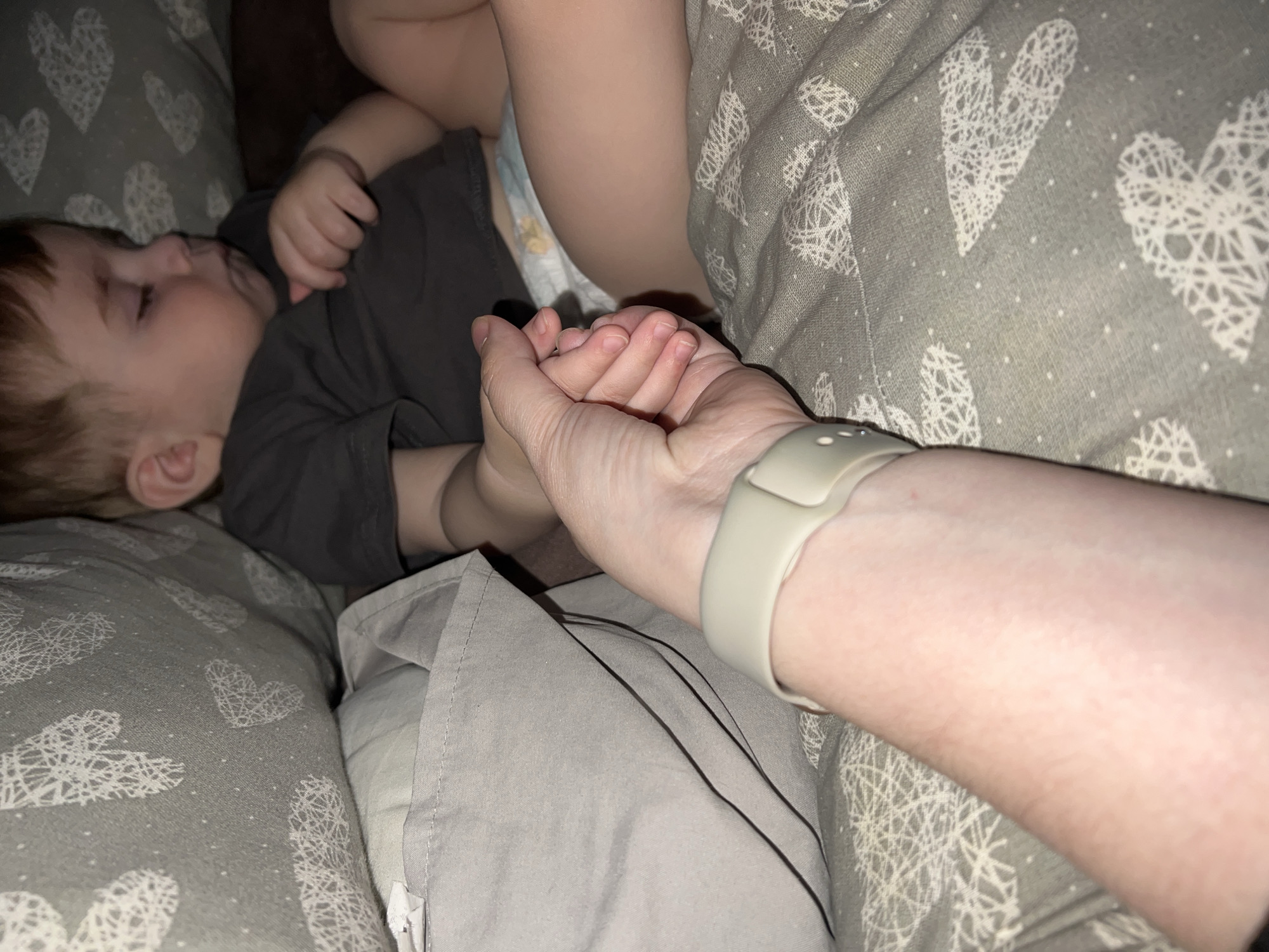 This close-up, dimly lit image captures a tender moment between an adult and a young child in an indoor setting, possibly in Noviy, Russia. The child, with reddish-brown hair, is lying on their side, seemingly resting or sleeping. They are dressed in a dark grey t-shirt, with a patterned diaper visible. An adult's arm extends into the frame from the right, gently holding the child's small hand. The adult wears a light beige or off-white smartwatch or fitness tracker on their wrist.
The subjects are surrounded by soft furnishings. A prominent grey pillow or blanket, adorned with numerous white, intricately patterned hearts and small white dots, is visible in the foreground and beneath the child. Another lighter grey fabric, possibly a blanket or part of the child's clothing, is seen closer to the adult's arm. A darker, solid-colored fabric, likely a couch or another piece of furniture, forms the background.
The lighting is soft and indirect, contributing to a calm and intimate atmosphere, suggestive of a quiet evening or a naptime moment. The image conveys a sense of comfort, security, and affectionate connection between the two individuals. No visible text is present in the image.
