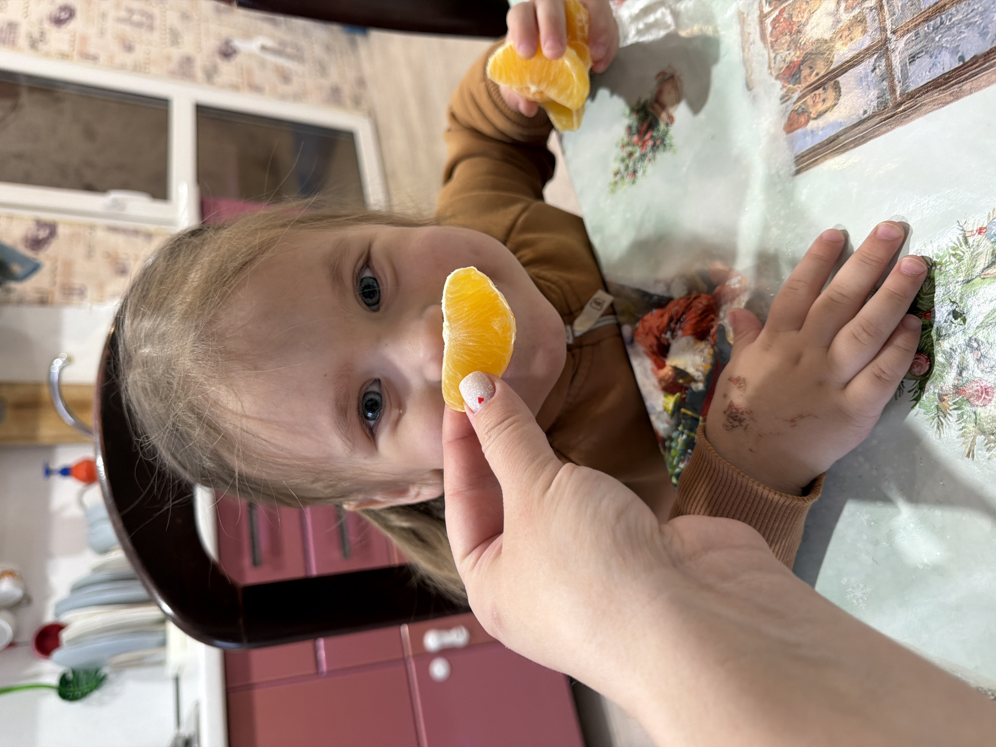 An indoor scene in Chaikovsky, Russia, depicts a young child seated at a table, interacting with an adult's hand. The child, with light brown hair and blue eyes, wears a brown long-sleeved garment and displays a smile. An adult's left hand, featuring a white manicured thumbnail with a red heart design, offers a peeled orange segment to the child. The child's left hand concurrently holds additional orange segments. The back of the child's right hand exhibits several small abrasions. The table is covered by a light green tablecloth patterned with winter or holiday-themed imagery, including snowflakes, evergreen branches, and a depiction of a window scene. Behind the child, a dark brown wooden chair back is visible. The background reveals a domestic kitchen or dining area with reddish-brown lower cabinets, a sink faucet, white dishes stacked in a drying rack, and a red and blue liquid dispenser. A white-framed glass door or window is also present against patterned wallpaper.