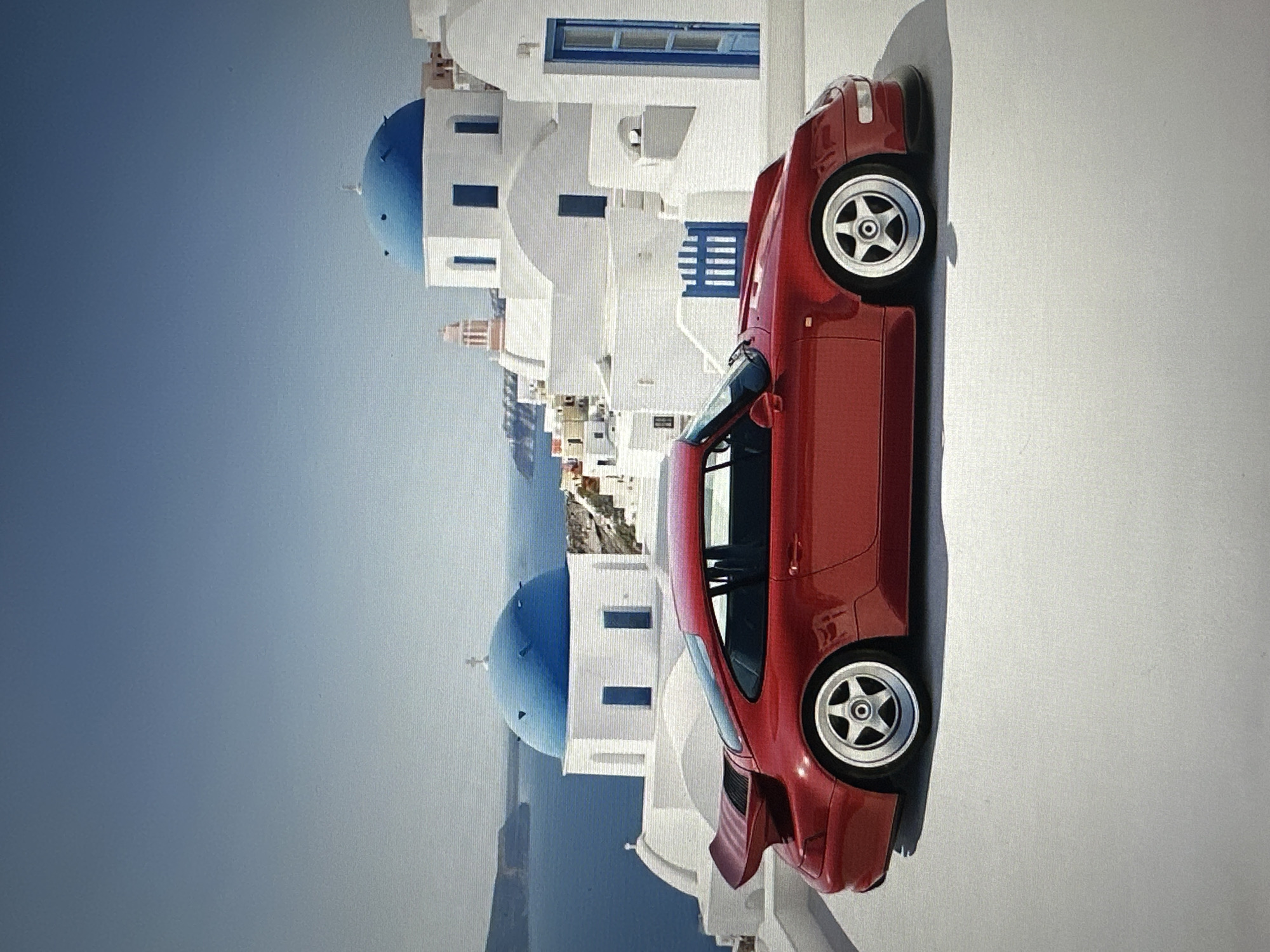 A vibrant red Porsche 911 coupe is parked on a white, flat surface with a backdrop of white buildings with blue domes and blue doors, characteristic of a Greek island. The sea and distant islands are visible in the background under a clear blue sky. The car is angled slightly towards the viewer, showcasing its sleek design and iconic spoiler. This image is not of Tallinn, Estonia.