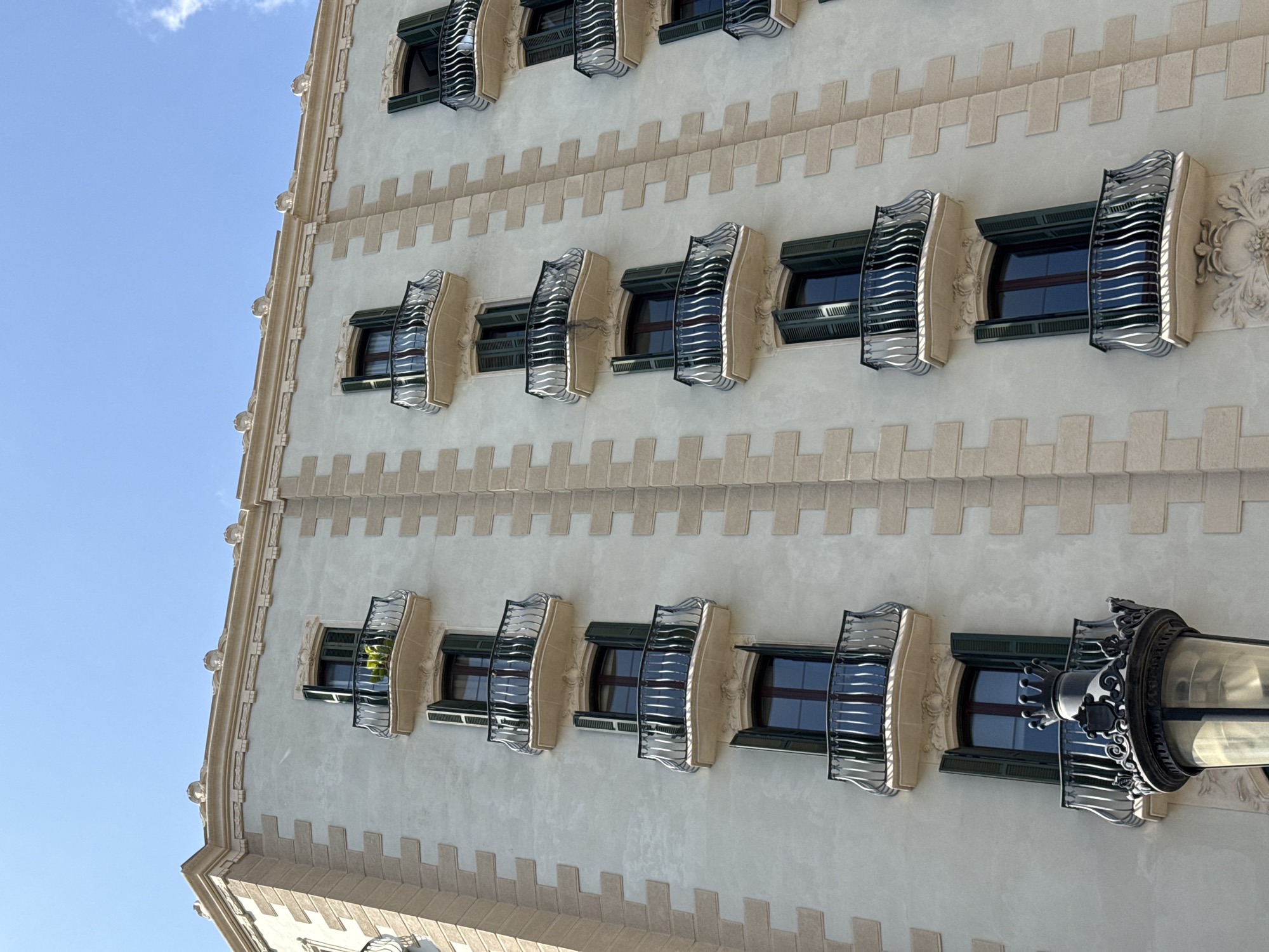 A multi-story building façade, characteristic of Barcelona, Spain, is captured from a low-angle perspective under a clear blue sky with minimal cloud cover. The building features a pale gray stucco finish contrasted by light tan, rusticated stone quoins that delineate vertical sections and corners.
The façade presents five visible vertical columns of windows, each equipped with dark green louvered shutters, mostly open, and a projecting semicircular balcony. These balconies are supported by decorative stone corbels and feature ornate black wrought-iron railings with curved vertical bars. Reflections of the sky and surroundings are visible on the windowpanes and the metallic railings. One balcony on the upper left side contains a small green potted plant.
A detailed cornice crowns the building, adorned with light-colored, sculpted busts or faces spaced along its length. In the lower-left foreground, a substantial black metal street lamp, partially visible, exhibits intricate decorative elements including a crown-like finial and a shield crest. Another ornate stone relief is visible below a window on the lower right section of the building. No people are visible in the scene.
