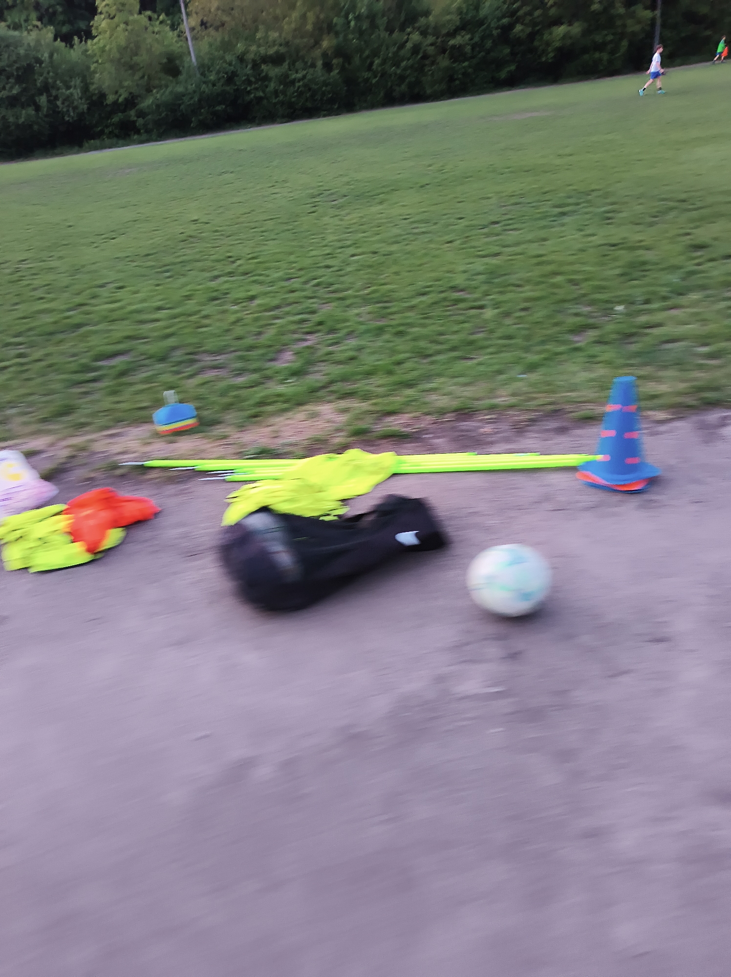 The image depicts a soccer field setting. In the foreground, a collection of training equipment is visible, including a soccer ball, a black bag, yellow poles, blue and orange cones, and training vests. The background shows a grassy field, with two individuals in the distance, possibly engaged in physical activity. The lighting suggests either late afternoon or early evening. Without specific landmarks, confirming the location as Ternopil, Ukraine, is not possible solely from visual data.