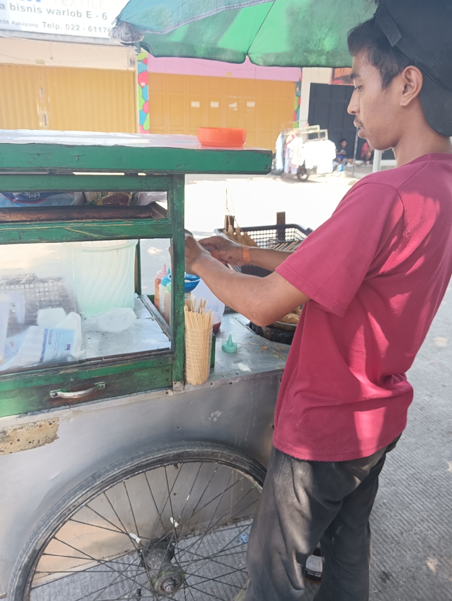 In Gandasari, Indonesia, a person wearing a maroon t-shirt and black pants stands behind a food cart. The cart has a green frame and a glass display case. Inside the display case, there are containers of liquid, possibly drinks, and what appears to be ice. Several bottles of condiments, like ketchup and chili sauce, are visible, along with a container of wooden skewers. The person is in the process of preparing food, holding a funnel and a bottle. The cart has a large wheel at the bottom. In the background, there are buildings with yellow and pink shutters, suggesting a commercial area. The scene appears to be taking place outdoors, on a paved surface.
