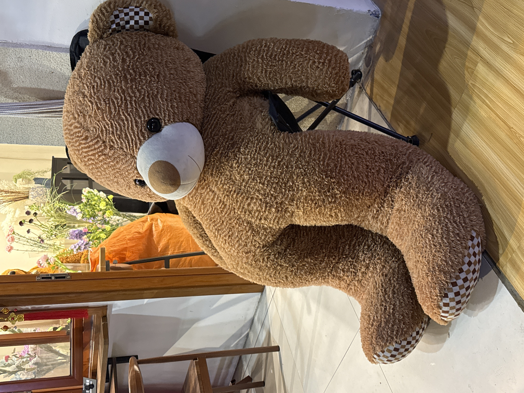 A large, brown teddy bear sits on a chair in an interior space. The bear has a checkered pattern on its ears and paws. In the background, there is a wooden cabinet topped with an arrangement of flowers and an orange cloth. The presence of Chinese-style decorations suggests the location is likely Qianjiang, China. The floor is a combination of tile and wooden planks.