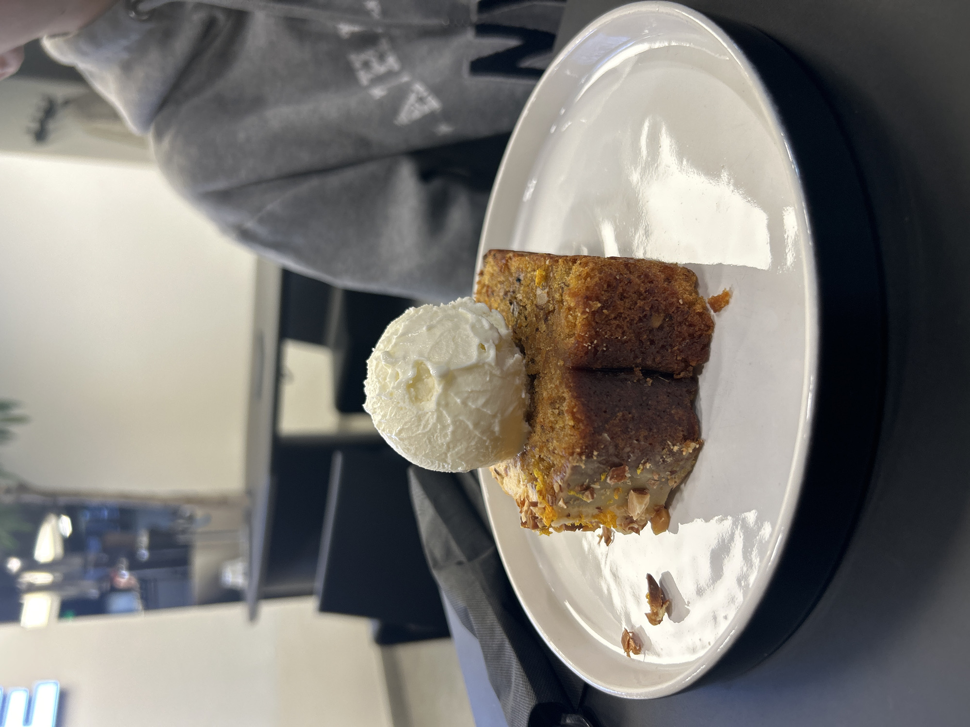 A plate with a slice of cake and a scoop of ice cream sits on a table, likely in a cafe or restaurant. The cake appears to be a moist, dense variety, possibly carrot or spice cake, with visible nuts or other ingredients. A scoop of white ice cream or whipped cream rests alongside the cake. The plate is white with a dark rim. The background shows a person wearing a grey hoodie. The specific location in Izhevsk, Russia, cannot be determined.