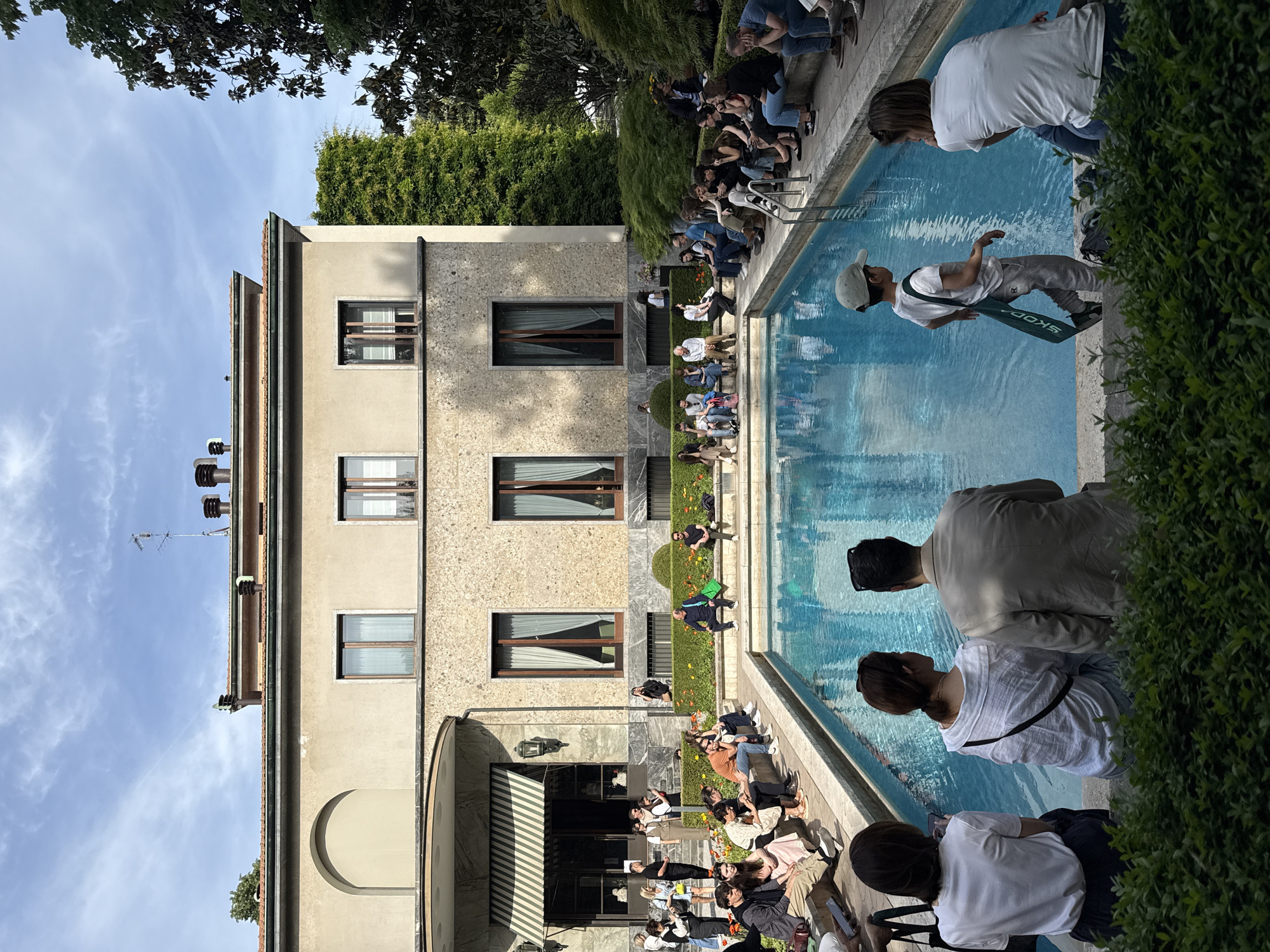 The image depicts a scene in Milan, Italy, with a body of water - either a pool or a large fountain - in the foreground. A modern building is on the left side of the image, with several rectangular windows. The building's facade appears to have a rough, textured finish. A crowd of people surrounds the water's edge. Some are sitting on benches or the edge of the pool/fountain, while others stand and interact with the water. One person seems to be either falling into the water or preparing to enter it. Green bushes line part of the water's edge. The sky is visible in the upper-left of the image. The overall light suggests it is daytime. The image is rotated counterclockwise.