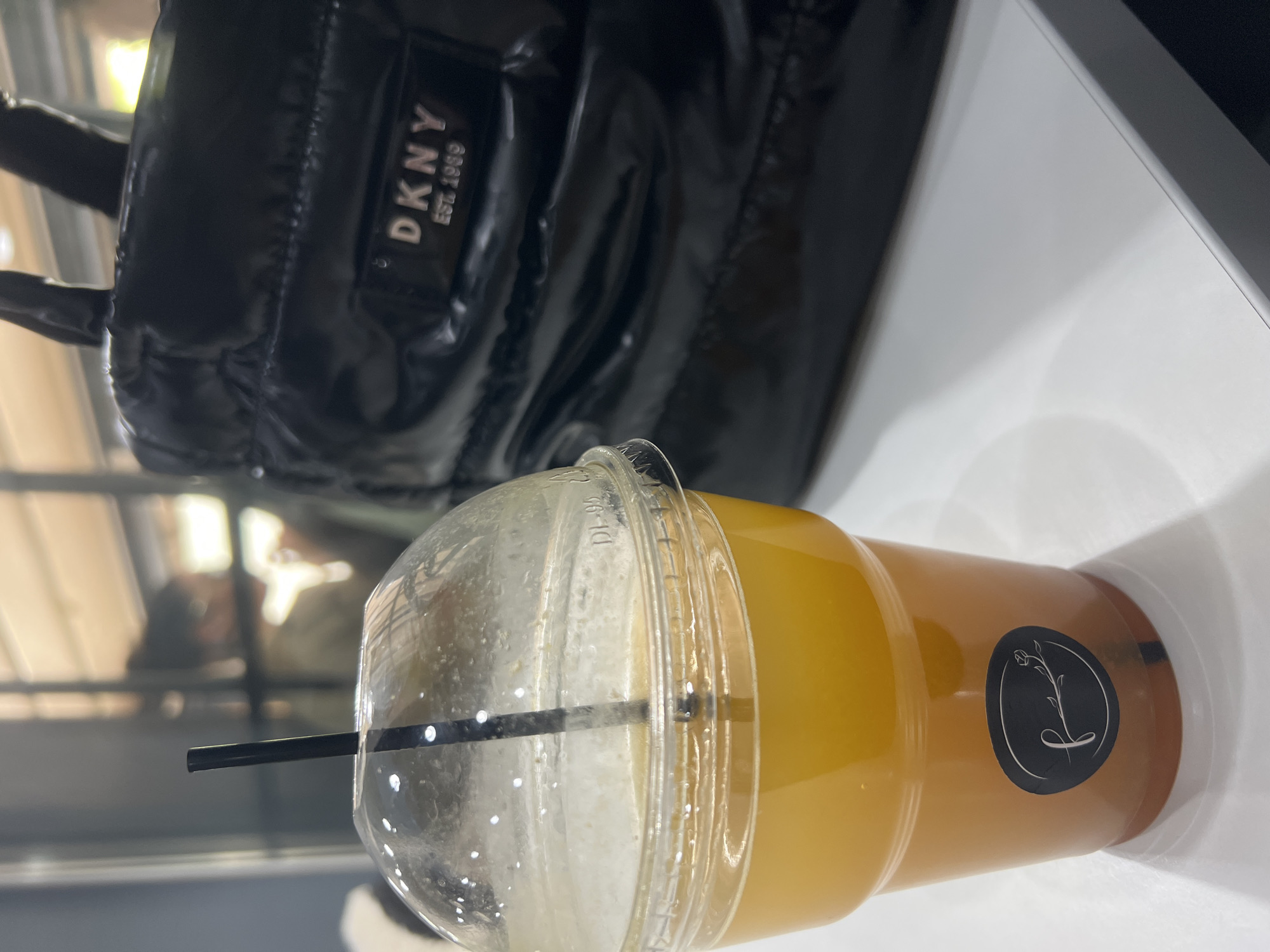 The image shows a plastic cup with a yellow beverage and a black straw, capped with a clear plastic dome lid. The cup has a round black logo with a stylized white flower on the side. The cup is placed on a white surface. In the background, a black DKNY bag is partially visible. The scene appears to be indoors, with a blurred view of an outdoor area through a window. While the origin of the image is not clear, it is impossible to confirm that it was taken in Plovdiv, Bulgaria without more information.