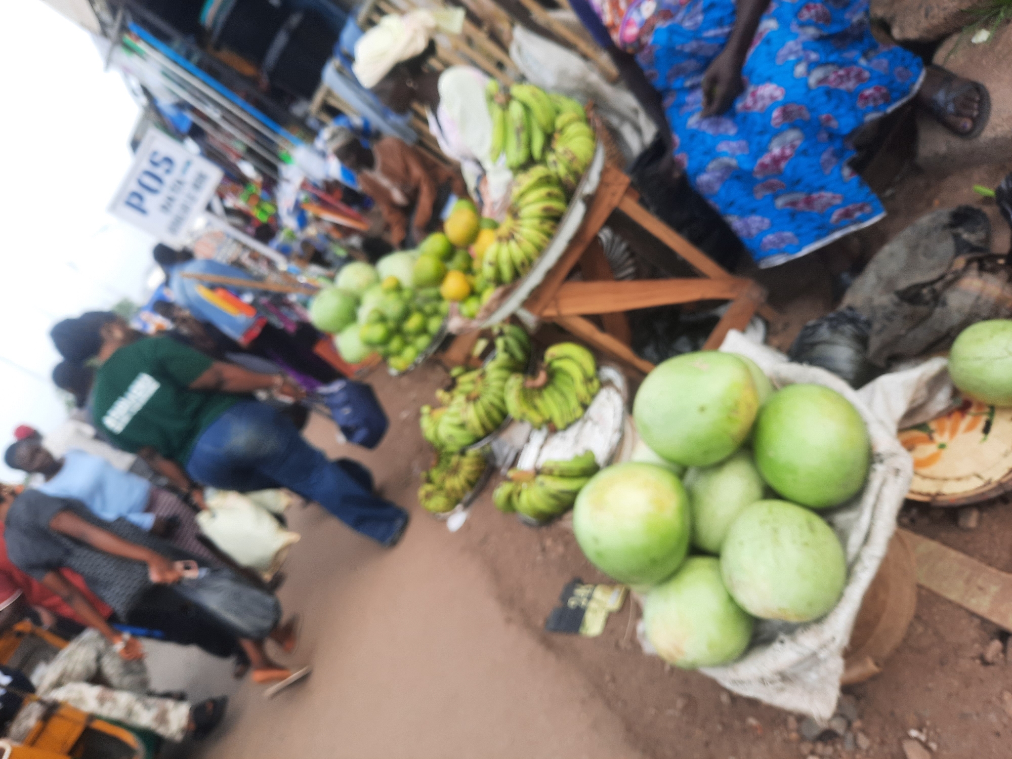 The image shows a blurry market scene in what appears to be Jos, Nigeria. Several people are visible, including vendors and potential customers. A woman in a blue patterned dress is sitting, likely selling goods. Fruits, including bananas, mangoes, and watermelons, are displayed on tables for sale. A sign with "POS" is present, indicating Point of Sale services. Other people are seen walking around or interacting with the vendors. A yellow tricycle can be seen on the left side of the picture. The overall setting suggests a busy marketplace in the city.