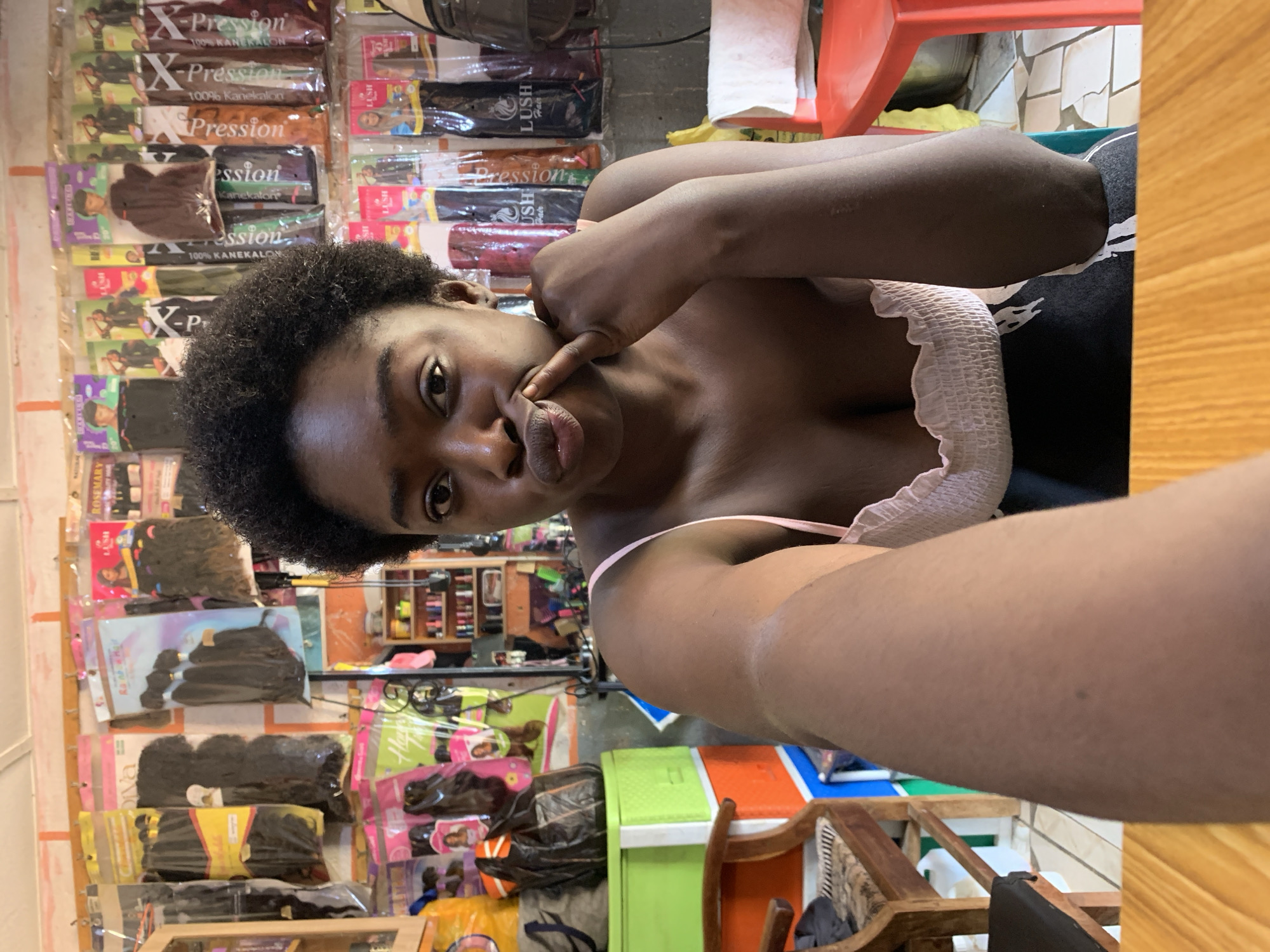 A person is taking a selfie indoors. The person has dark skin and short, curly black hair. The person is wearing a light-colored top with a ruffled edge. The person's right index finger is touching their upper lip and nose, as if pushing the nose upwards.
In the background, there is a wall covered with hair product packaging. Some packages are labeled "X-Pression" and "Lush Hair." Below the wall, there are various items on shelves and the floor, including packaged hair extensions, containers, and chairs.
The picture appears to be taken in a shop or store in Rafin Par, Nigeria that sells hair products.