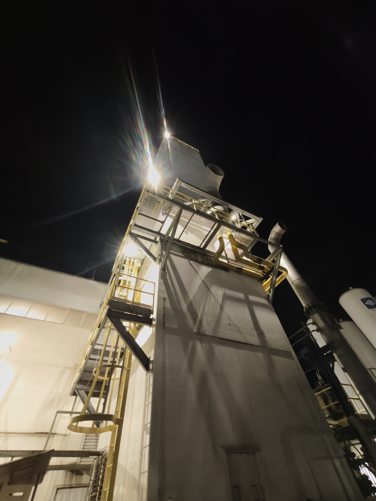 At night, a tall, white industrial structure dominates the view. The structure features metal walkways and a ladder on its exterior, illuminated by a bright light at its upper section, creating a lens flare effect in the dark sky. Adjacent to the main structure are pipes and cylindrical tanks, also painted white. The scene is set against a completely dark sky, with only the structures and their lights providing illumination. The location is Unknown, Unknown.