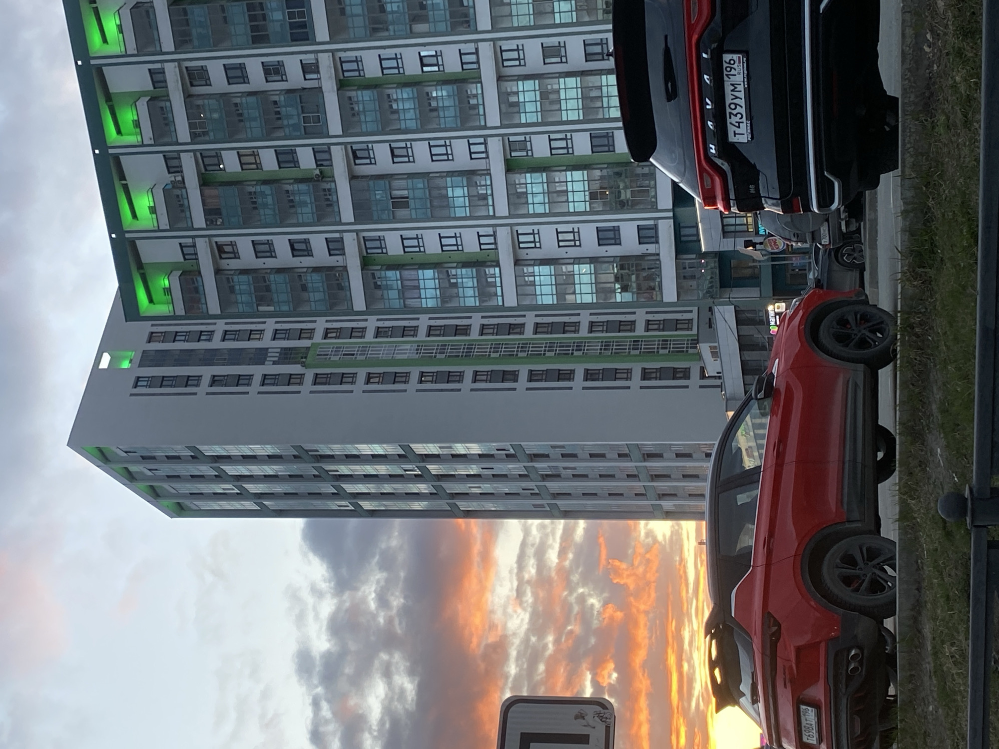The image shows a tall, multi-story building in Yekaterinburg, Russia, as seen from ground level. The building has a modern facade with many windows and green accents. A black SUV with the license plate "T439YM 196 RUS" and a red SUV with the license plate "T698AT 196 RUS" are parked in front of the building. The sky above the building displays a cloudy sunset with orange and gray tones. A street sign is also visible in the lower part of the frame.