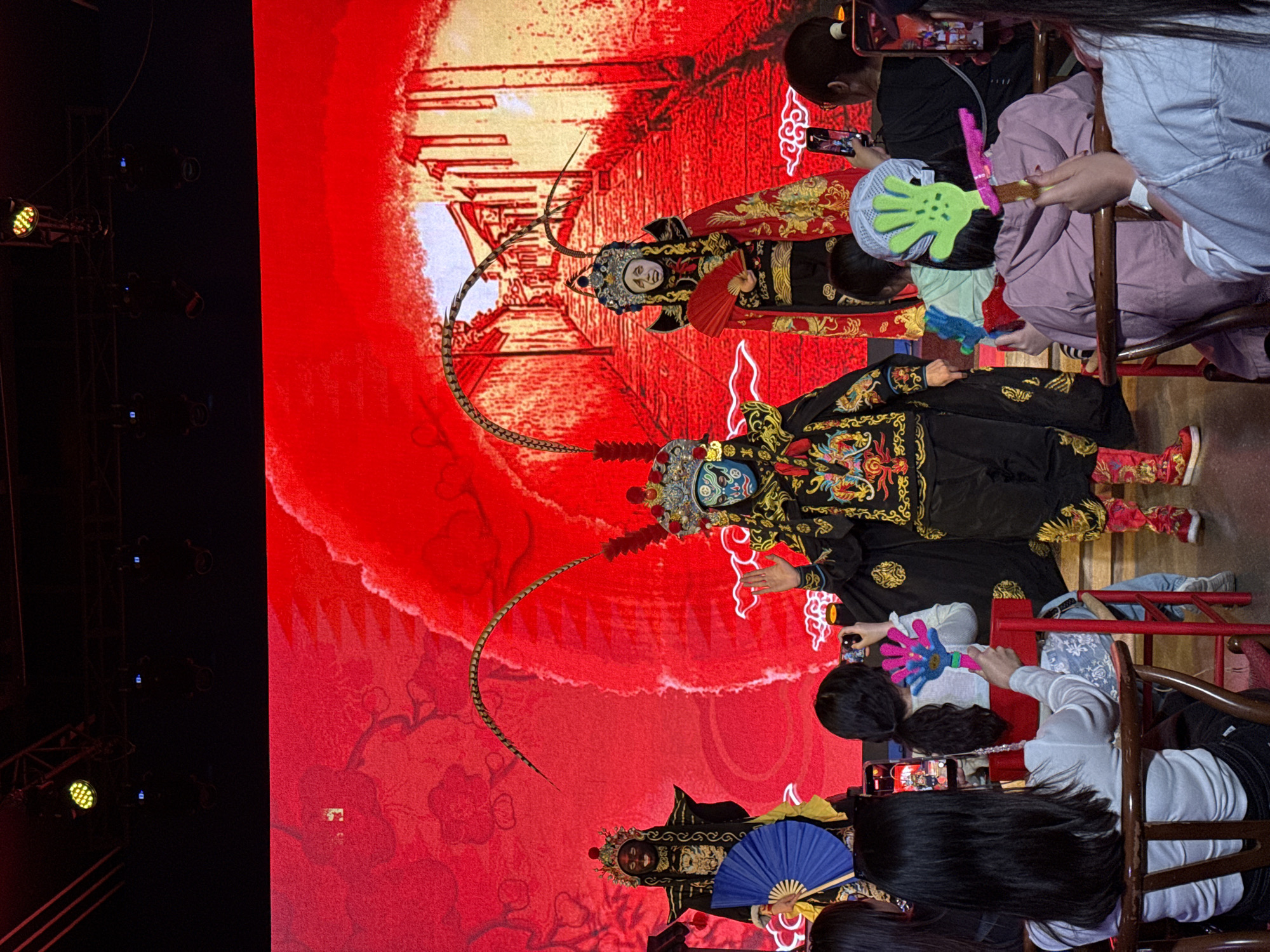 The image depicts a performance, likely a Chinese opera or similar theatrical event, taking place in Yuzhong District, China. Two performers in elaborate traditional costumes are on stage in front of a large red backdrop. They wear ornate headdresses and colorful makeup, and their costumes feature intricate embroidery.
Several audience members are seated in the foreground. Many are holding up phones, suggesting they are taking photos or videos of the performance. Some audience members are also holding fans. The background is a mix of the stage setting and the theater environment. The lighting indicates a stage production setting.