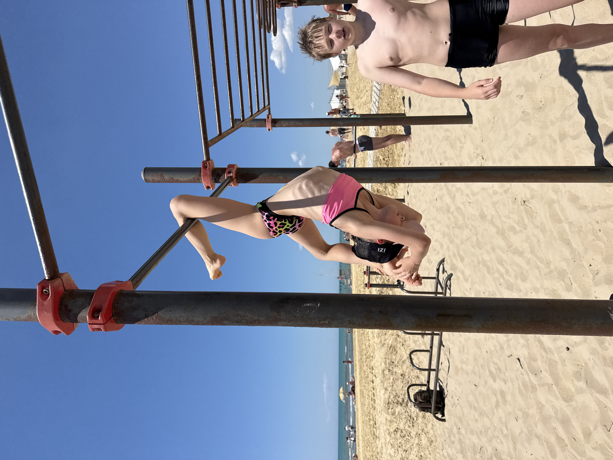 A young person is upside down on horizontal bars at a beach in Natukhayevskaya, Russia. The person is wearing a leopard print and pink bikini and a black cap. Another young person, a boy, stands to the right of the bars, facing the camera. In the background, the sea, beach, and other people can be seen. The weather is sunny with a clear blue sky.