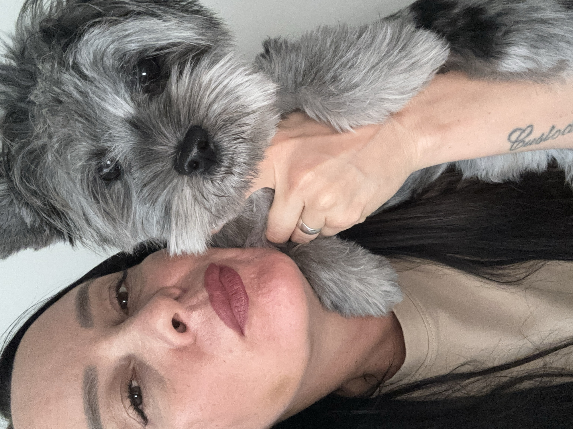 A close-up, intimate portrait captures a woman and her small dog indoors. The image is oriented such that the woman's head and upper torso occupy the lower portion of the frame, with the dog positioned above her, resting on her.
The woman has long, dark hair visible around her shoulders and against a plain, light background, likely a wall. She wears a light beige or tan t-shirt. Her face features defined dark eyebrows, subtle eye makeup, and a muted reddish-brown lipstick. Her gaze is directed towards the camera. A silver band ring is visible on the ring finger of her right hand, which gently rests on the dog's fur near its front paw. On her right forearm, a stylized script tattoo, possibly a name like "Gustavo" or "Custodio," is partially visible.
The dog, a fluffy breed resembling a Schnauzer with mottled grey and black fur, has its face intimately close to the woman's. Its dark, expressive eyes look directly at the camera, and its dark nose is prominent. The interaction suggests a close bond and affection between the owner and her pet.
The setting appears to be a domestic indoor environment, characterized by a simple, uncluttered white background. The soft lighting suggests a daytime setting. This moment of companionship takes place in Chaikovsky, Russia.