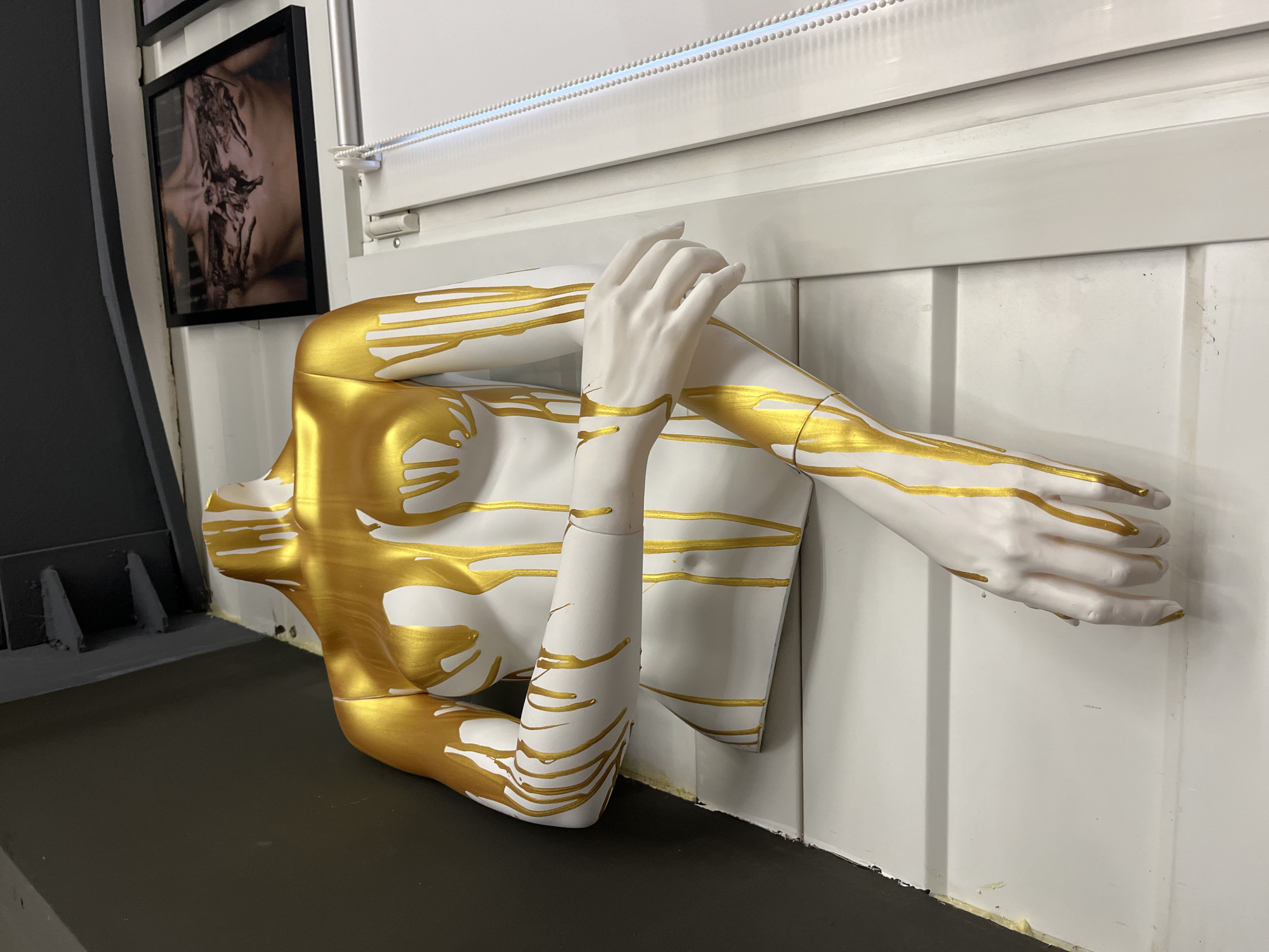 A half-mannequin painted white with gold streaks lies on a black surface, possibly a display or shelf, pressed against a white, horizontally paneled wall. The mannequin is cut off mid-torso and features two arms, one bent with the hand resting near the shoulder, the other outstretched. The gold streaks appear like dripping paint or metallic embellishments. Above the mannequin, a string of small, spherical lights runs horizontally along the top of the wall. To the left, partially visible, is a black-framed picture and a gray surface or wall. The location is Chaikovsky, Russia.