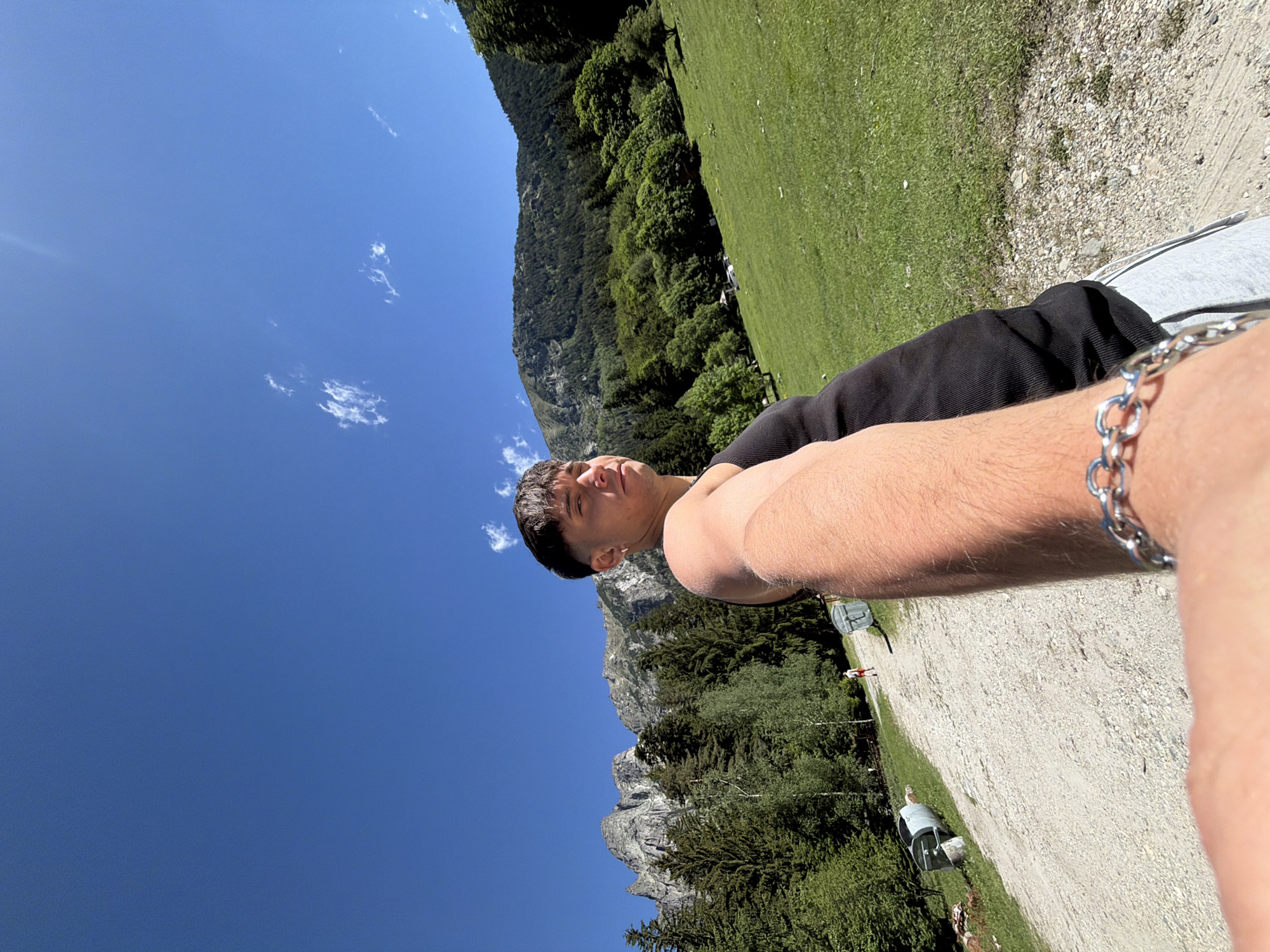 The image is a selfie taken outdoors in a mountainous, grassy environment under a clear blue sky with scattered clouds. In the foreground, a young man with short dark hair is captured from the chest up, wearing a black tank top and a silver chain bracelet on his left wrist. He is looking towards the right side of the frame. Behind him, a gravel path stretches into the distance. To the left of the path, large, rocky mountains are visible, interspersed with dense green pine trees. To the right of the path, a lush green meadow slopes upwards towards a forested mountainside. A person in red shorts is walking away from the camera on the path in the middle ground. Two large, cylindrical metal containers, possibly for waste disposal, are situated beside the path. The overall scene suggests a rural or natural setting, potentially near a hiking trail or a natural park. The location is identified as Govedartsi, Bulgaria.