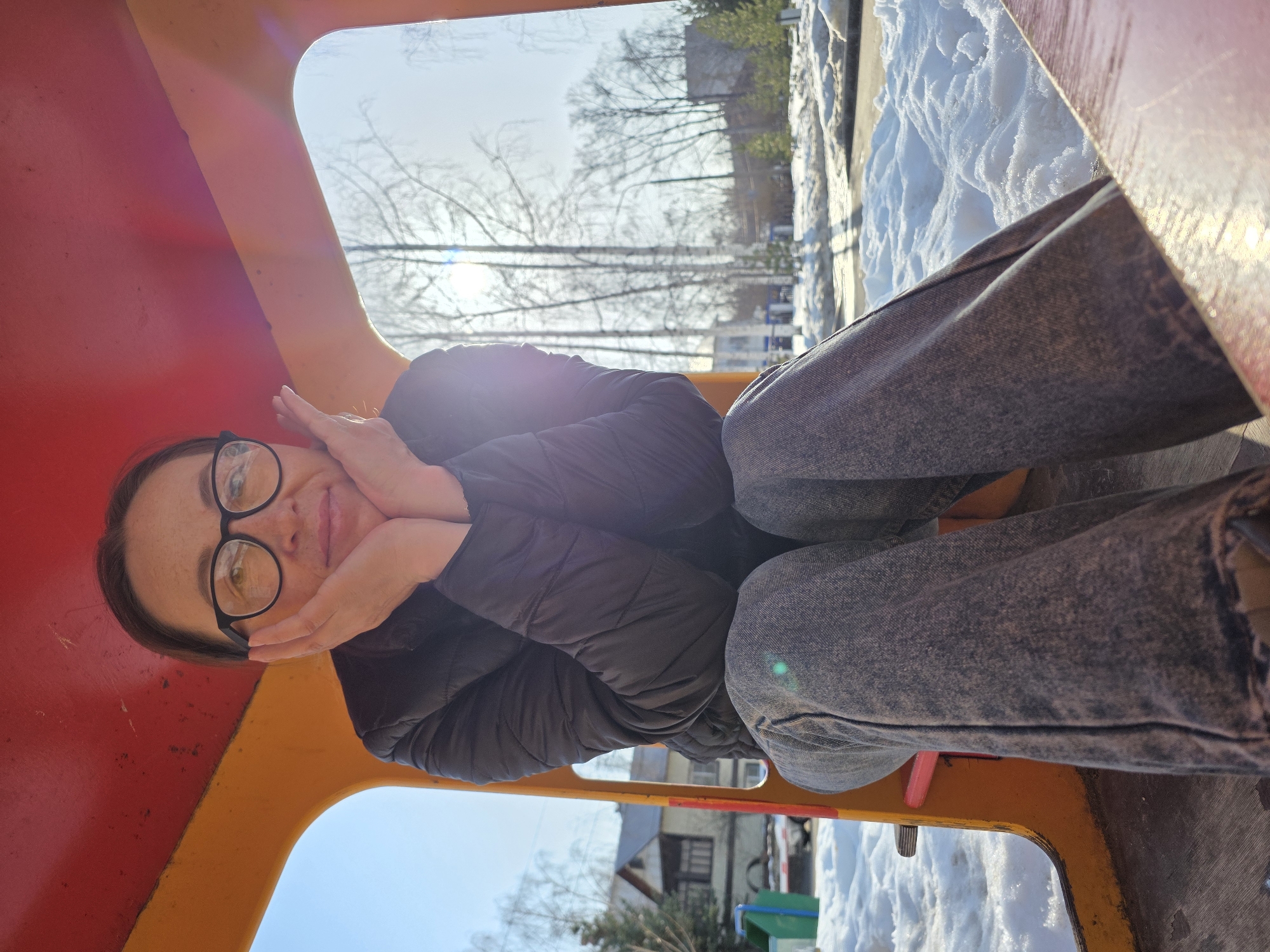A woman is depicted sitting inside a brightly colored playground structure, likely in a park or residential area in Chaikovsky, Russia. She wears a black puffer jacket, dark wash jeans, and black-framed eyeglasses, with her hands resting against her cheeks, looking directly at the camera with a subtle smile. The interior of the structure features a red ceiling and orange-painted walls/frame, with a wooden floor where she is seated. Outside the structure, patches of snow cover the ground, indicating a winter or early spring season. Bare deciduous trees are visible in the midground against a clear, bright sky, with strong sun glare emanating from the upper right, partially obscuring a building and other distant structures. A residential-style building with a pointed roof is visible to the left, and a green trash receptacle stands nearby in the snow.