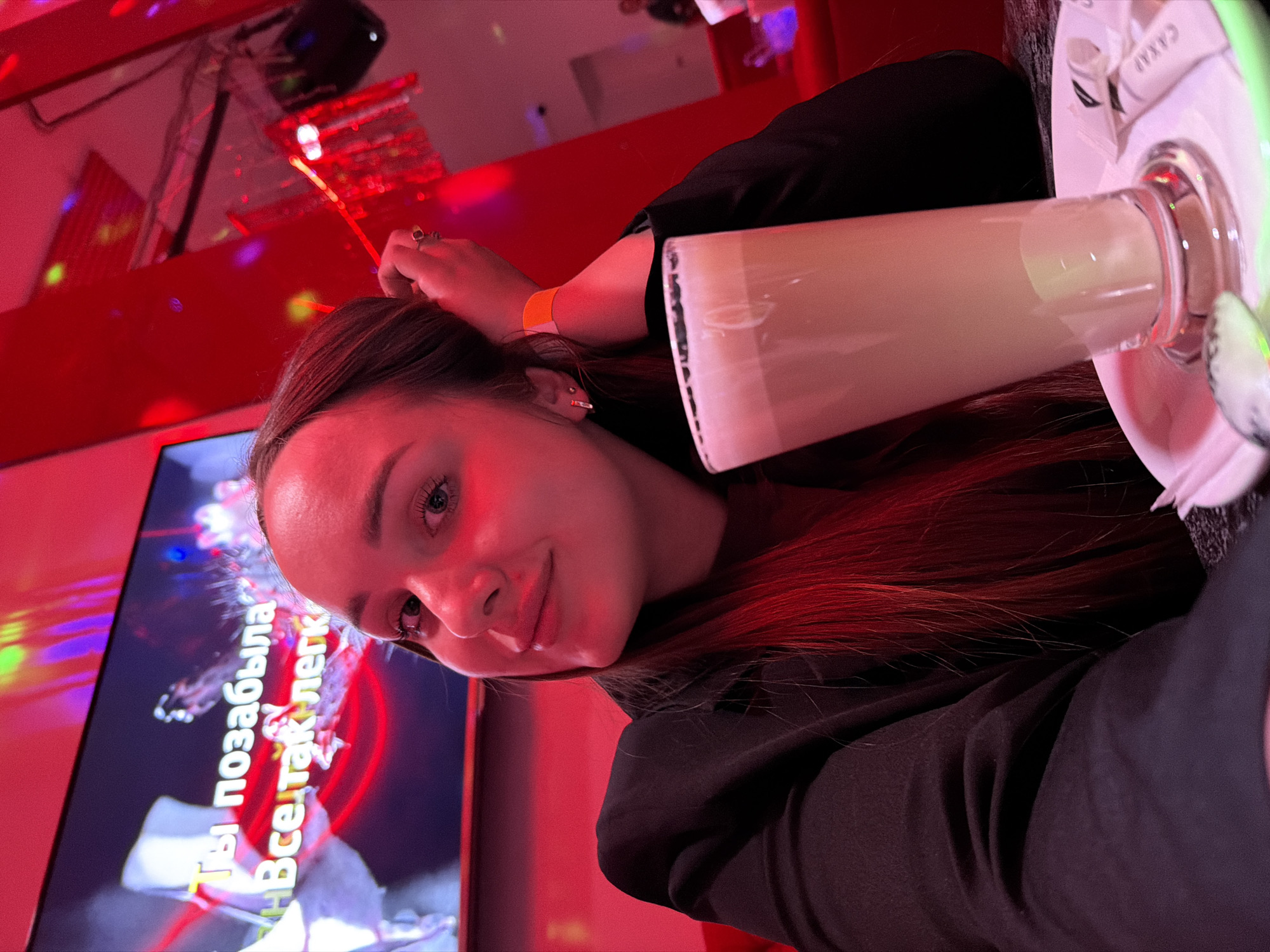 A young woman with auburn hair and fair skin poses for a selfie in a karaoke bar, as indicated by the karaoke screen in the background that displays Russian text, and a disco light. She is holding a tall glass of a white foamy liquid, possibly a cocktail. The environment is illuminated with red lighting. She is wearing a black jacket and orange wristband. A ring is visible on her right hand. A coaster and possibly a napkin with the word "Сахар" are visible near the glass. The location could be Chaikovsky, Russia, based on the Cyrillic text on the karaoke screen.