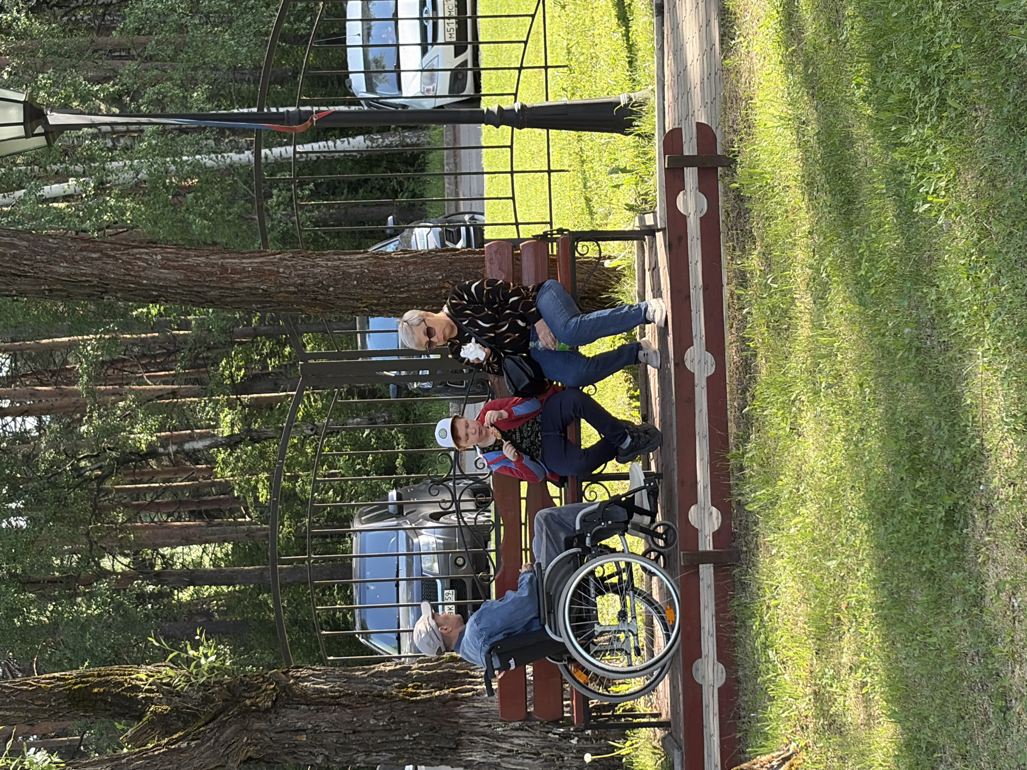 In the city of Chaikovsky, Russia, a woman in a patterned top and jeans sits on a park bench next to a boy in a red and blue jacket. An elderly man in a denim shirt and cap is in a wheelchair on the same bench. They are positioned in front of a metal fence and a row of parked cars. A lamppost stands nearby. The environment includes trees and grass.