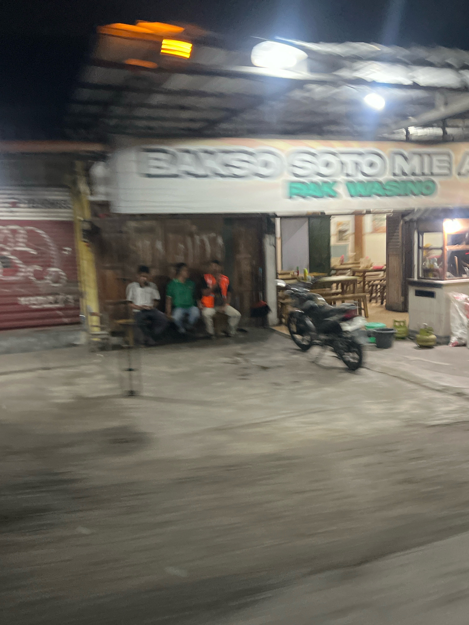The image shows a blurry outdoor scene at night, likely in front of a food establishment. A sign above reads "BAKSO SOTO MIE A PAK WASINO," indicating the name of the business. In the foreground, a black motorcycle is parked. Several people are seated on a bench to the left, one in a white shirt, another in a green shirt, and a third in an orange vest. Behind them, tables and chairs are visible, suggesting a dining area. The overall environment appears to be a street or an open-air dining space. There are gas tanks and a bucket near the motorcycle.