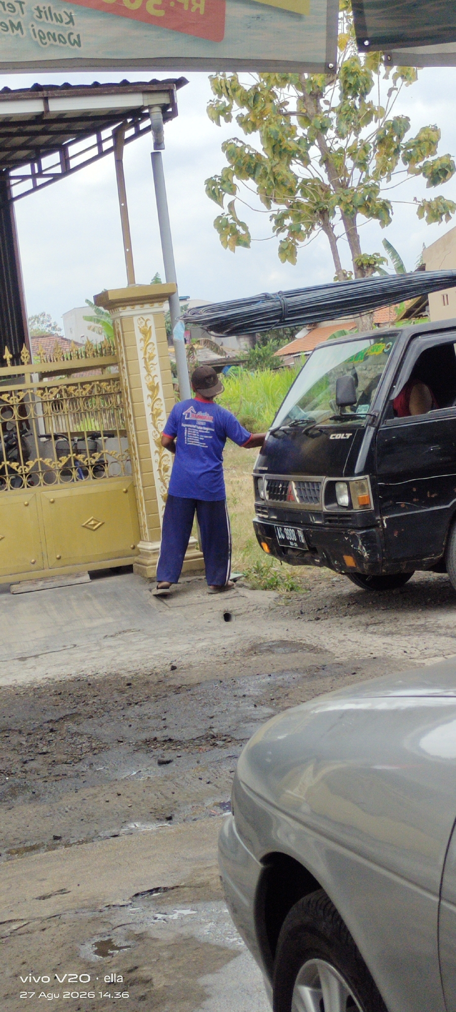 In Simo, Indonesia, a person wearing a blue t-shirt and blue pants with white stripes, and a brown hat, stands on a dirt and concrete surface next to a gold-colored gate and a black Mitsubishi Colt pickup truck. The person is facing the truck and appears to be reaching towards its front, possibly interacting with it. The truck's license plate is visible and reads "LC 9898 PN". Behind the gate, there is a building with a metal roof structure. A tree with light green and yellowing leaves is in the background, along with other residential buildings and green vegetation. The sky is overcast. In the foreground, the hood and fender of a silver car are visible.
