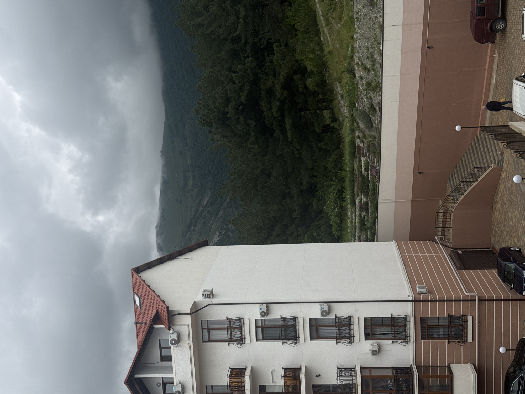 Here is a detailed description of the image:
The image shows a building with a light-colored facade and a red-tiled roof. The building appears to have multiple floors, with windows and small balconies on each floor. Each window has metal bars. Air conditioning units are mounted beneath some of the windows.
The building is adjacent to a hillside covered in dense vegetation, partially obscured by fog. The weather appears cloudy.
To the right of the building is a wall, painted in a light brown color with white strips and with a set of stairs. A few parked cars are visible, and a person is walking on the sidewalk next to the building.
The image appears to be taken in a mountainous region, possibly in Gelendzhik, Russia, known for its coastal location.