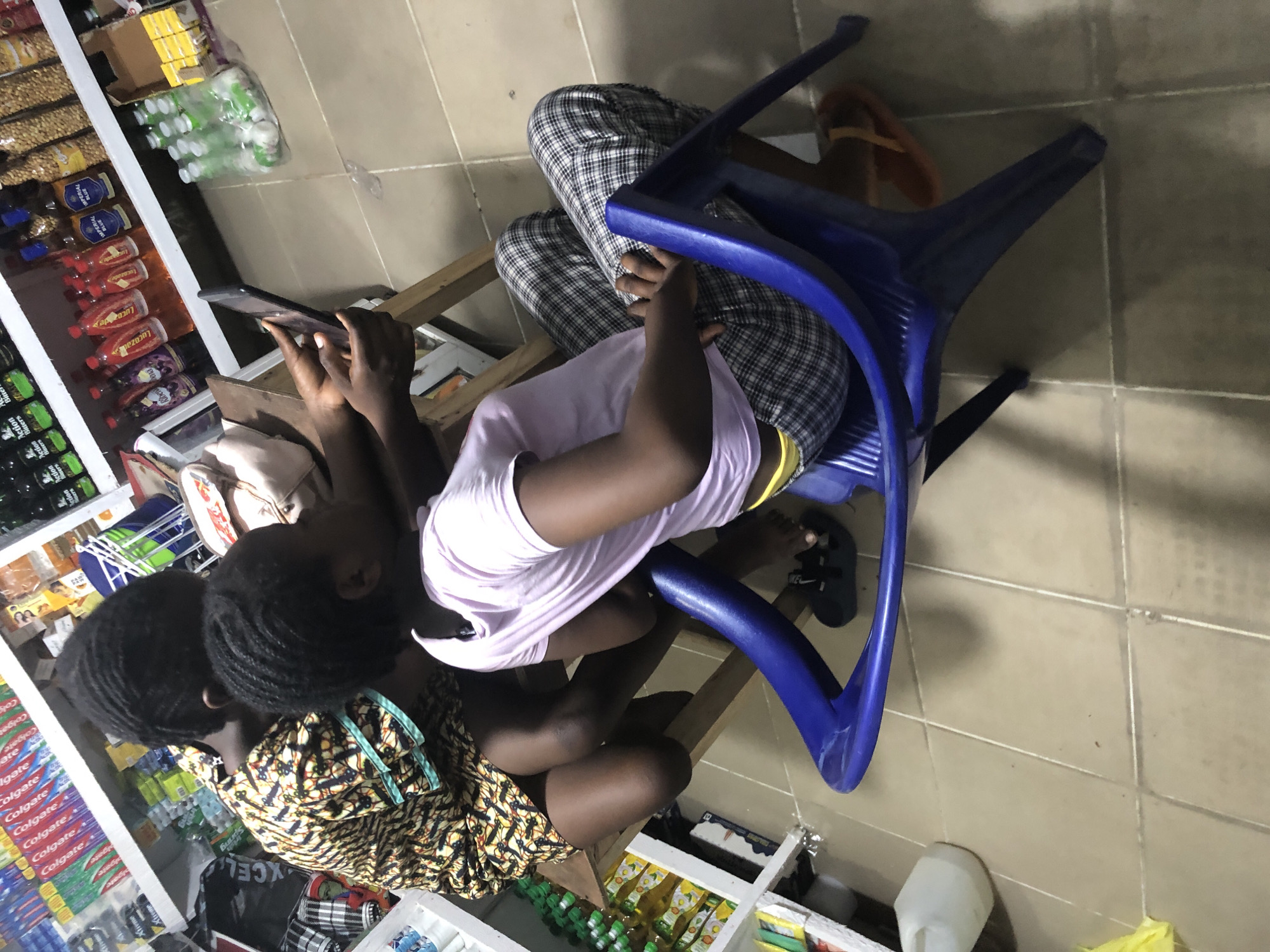 The image depicts an indoor scene, possibly a small shop or convenience store, in Ado, Nigeria. Two young individuals, appearing to be children or teenagers, are seated. One, wearing a patterned yellow and black garment, is seated on a wooden stool. The other, in a light purple t-shirt and checkered pants, is seated on a blue plastic chair. The individual on the chair is holding a smartphone and appears to be showing something on the screen to the person on the stool, who is looking at the device. Shelves stocked with various products, including what appear to be beverages and personal care items like toothpaste, line the background. The floor is tiled in a neutral tone. The overall lighting suggests an artificial indoor environment.