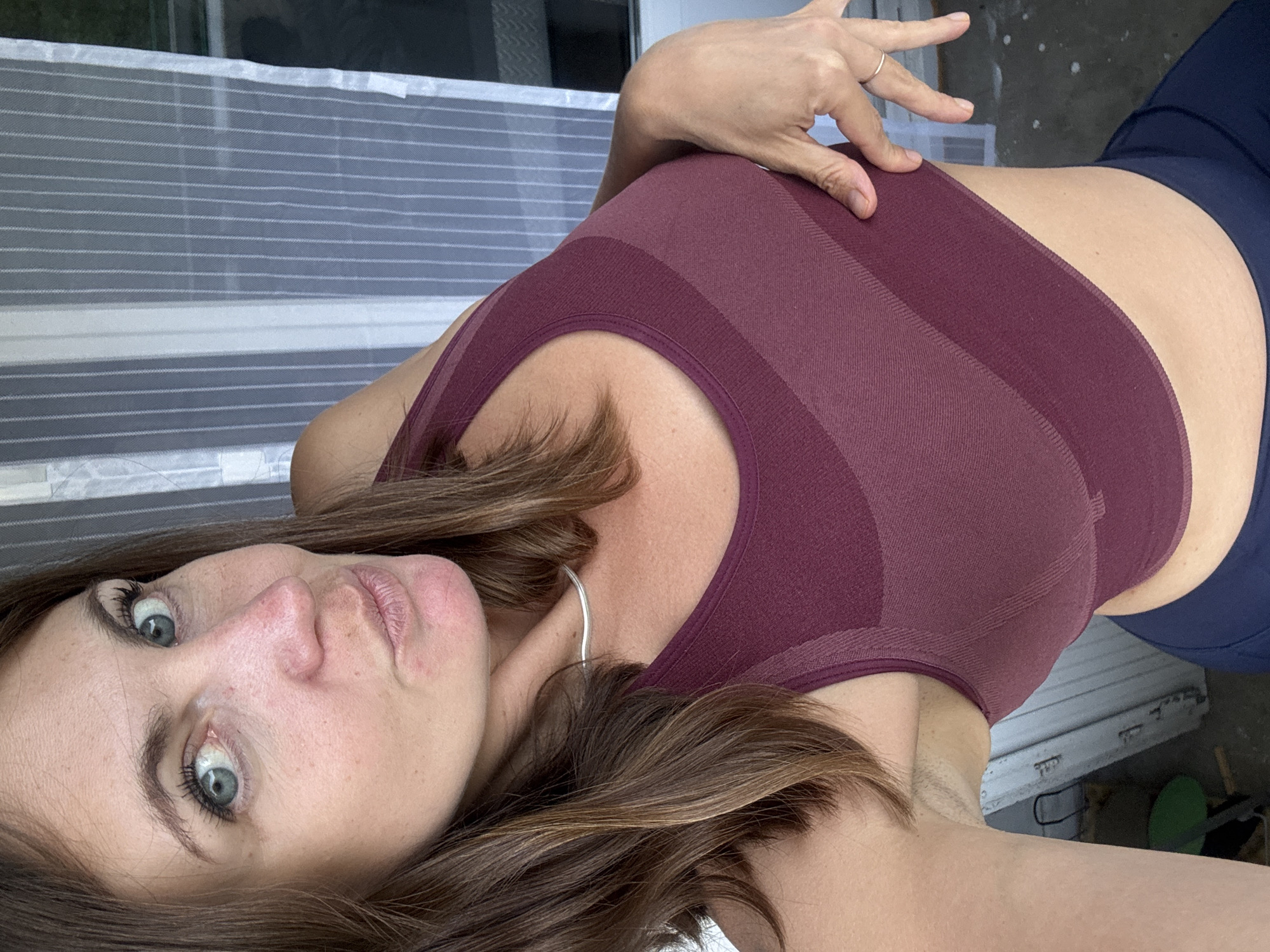 A woman with brown hair and blue eyes is posing for a photo. She is wearing a maroon sports bra and navy blue leggings. Her left hand is placed on her stomach, and she is looking at the camera with a playful expression. The background appears to be an outdoor setting, possibly a patio or balcony, with a striped curtain visible. There is no indication of the location being Saint-Claude, France.