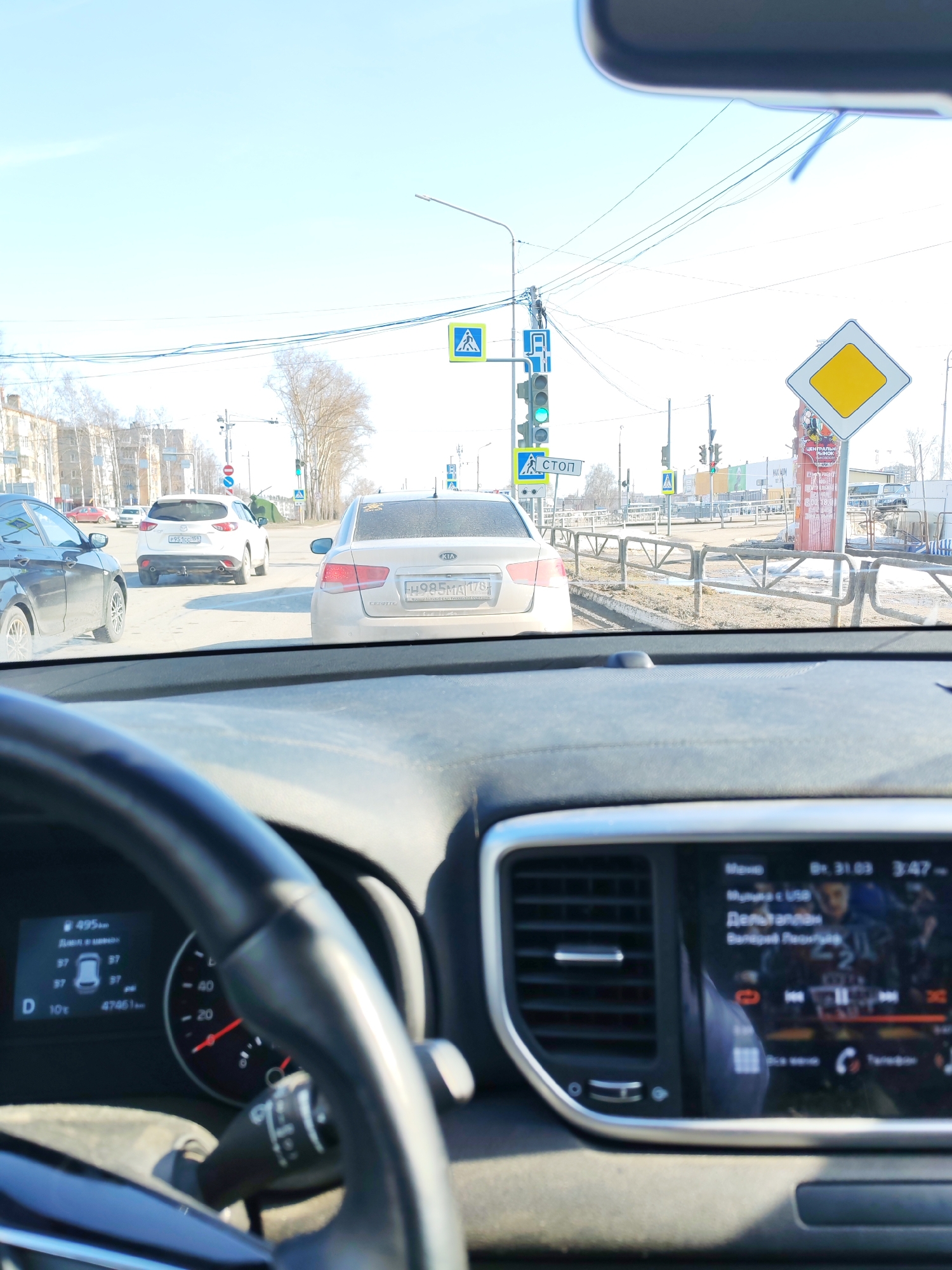 The image, taken from inside a car, shows a traffic scene in Chaikovsky, Russia. A Kia Cerato is directly ahead, with license plate H985MA 178. To the left, a white Mazda CX-5 with license plate P951CC 159 is visible. A traffic light showing green, along with a pedestrian crossing sign marked "Стоп" (Stop), is visible at an intersection. A yellow diamond-shaped road sign is also visible to the right. The car's dashboard shows a mileage of 47461 and a temperature of 10 degrees Celsius. The infotainment screen displays music information. The scene is set on a sunny day.