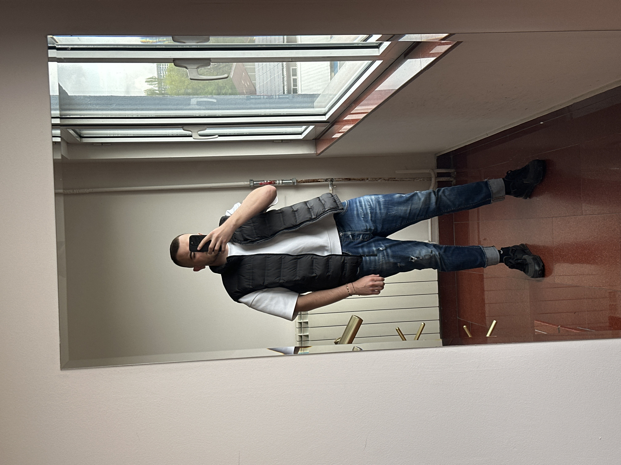A man is taking a mirror selfie in what appears to be an entryway or hallway. He is wearing jeans, a white t-shirt, and a black vest. He is holding a cell phone in his left hand, presumably to take the picture. The mirror reflects a room with light-colored walls, a tiled floor, and a window. Outside the window, a building and trees are visible. The location is not identifiable as Plovdiv, Bulgaria based on the given image.