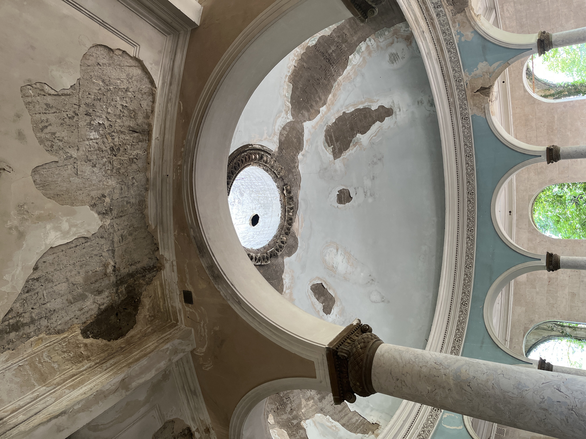 The image shows the interior of a dilapidated building, likely a former spa or sanatorium, in Tskaltubo, Georgia. The scene is characterized by decay and neglect. The ceiling exhibits significant water damage and peeling plaster, revealing the rough structure beneath. A central oculus in the dome allows natural light to enter, casting a stark illumination on the worn surfaces. Arched structures, supported by marble-like columns, frame views of arched doorways and windows, beyond which lush green foliage is visible, indicating an overgrown outdoor environment. Decorative moldings and painted elements, once ornate, are now faded and damaged. The overall impression is one of abandoned grandeur, with nature beginning to reclaim the man-made structure.