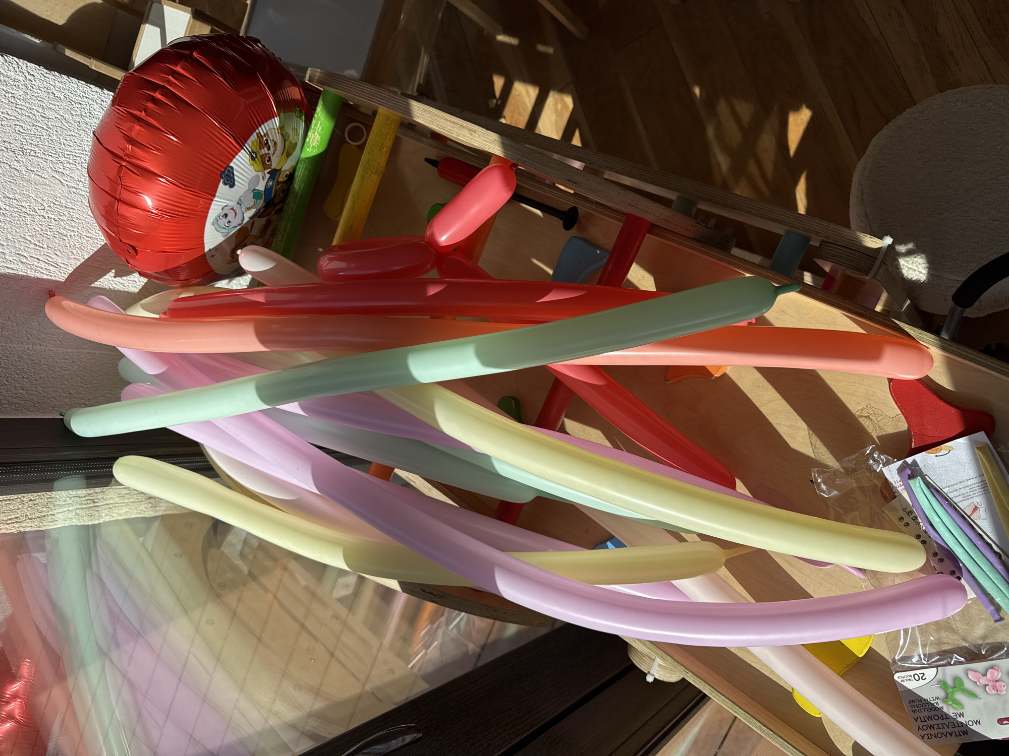 The image captures an indoor scene dominated by a pile of colorful, uninflated modeling balloons in pastel shades of pink, light green, light yellow, and purple. Resting on a wooden surface, these balloons suggest preparations for a celebration. A partially inflated, glossy red foil balloon featuring characters from Paw Patrol (including Chase, Marshall, Skye, Everest, and Rubble) is prominent on the left.
The balloons are situated on what appears to be a wooden play structure or furniture with visible slats. Strong, direct sunlight streams into the room, casting sharp, angular shadows from the wooden elements onto the floor and objects, indicating a sunny daytime. A white, textured wall is visible on the far left, and a fluffy, light-colored circular mat or rug is in the upper right background.
The presence of numerous uninflated balloons, a celebratory foil balloon, and a small hand pump (partially visible among the balloons) strongly implies preparations for a party or a festive event, likely a child's birthday given the Paw Patrol theme. Text on a plastic bag containing balloons on the right reveals "20 pcs" and "MODELING BALLOONS WITH PUMP", confirming their purpose. The scene evokes a mood of joyful anticipation for an upcoming event in Sofia, Bulgaria.