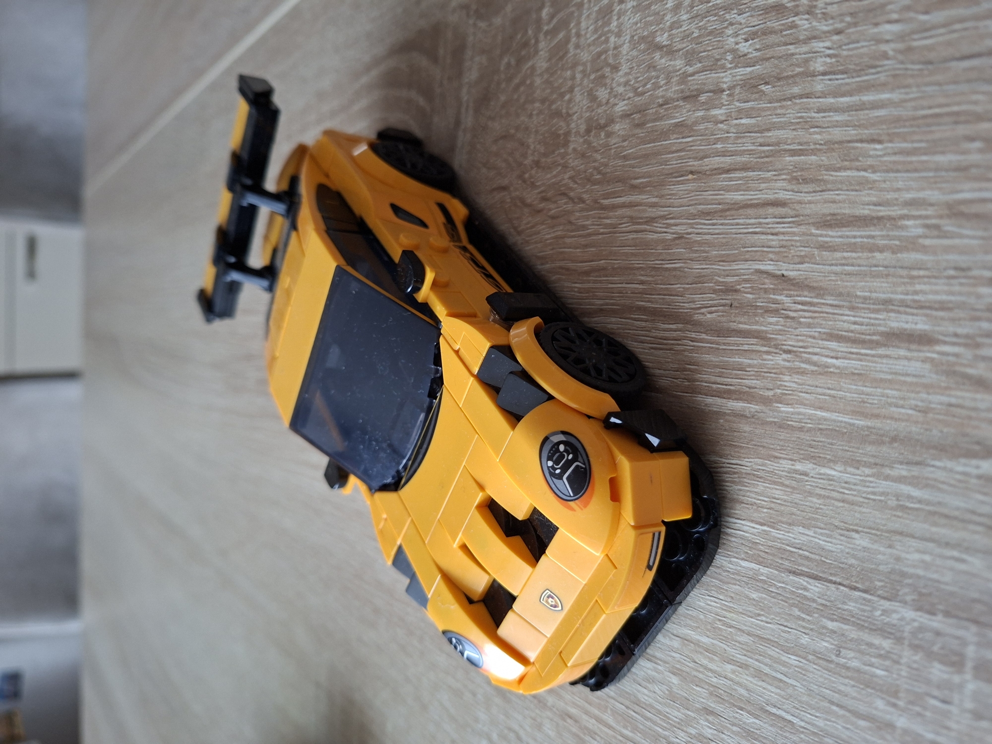 A yellow Lego toy car with a black spoiler is positioned on a wooden surface. The car is angled with its front to the left and its rear to the right. The background is blurred and appears to be an indoor setting with furniture. No people or specific landmarks of Baden-Baden, Germany, are visible.