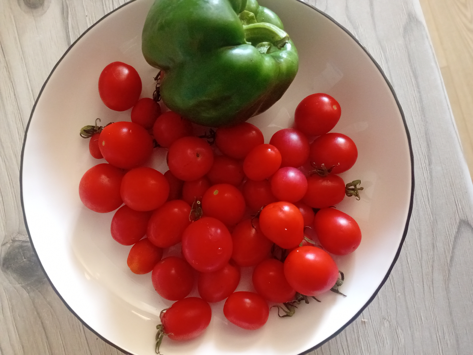 A white bowl with a black rim sits on a light-colored wooden table. The bowl contains numerous small, red cherry tomatoes and one large, green bell pepper. The tomatoes are scattered throughout the bowl, some clustered together, while the bell pepper is positioned to the right side of the bowl, taking up a significant portion of its space. The lighting suggests an indoor setting, possibly a kitchen or dining area. There are no people or animals present in the image, and no specific actions or interactions are depicted. The environment is a simple, uncluttered tabletop. The location in the city of Курманаевка, Russia, cannot be determined from the visual information in the image.