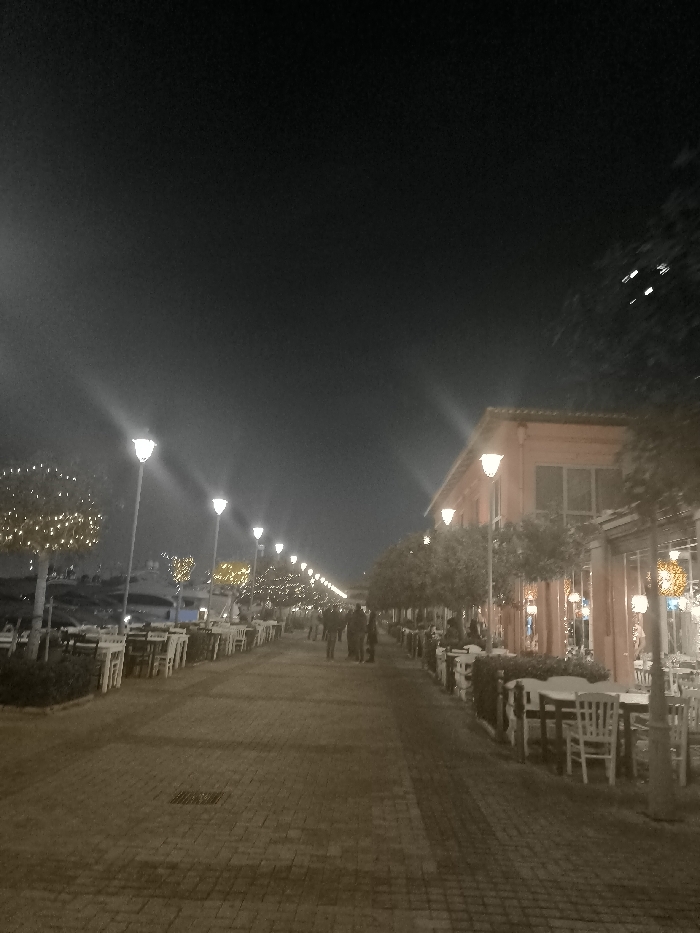 The image captures a vibrant nighttime scene of a pedestrian promenade in Palaio Faliro, a coastal suburb of Athens, Greece. A wide, paved walkway, appearing to be made of brick or stone pavers, stretches into the distance, illuminated by an array of lights.
On both sides of the promenade, numerous outdoor dining areas are set up with neatly arranged white tables and chairs, suggesting a lively row of restaurants, cafes, or bars. On the right, these establishments are housed within low-rise buildings characterized by a distinctive reddish-orange facade and large windows. Small, manicured trees line the walkway and stand in front of the buildings, many adorned with bright, decorative string lights that add a festive glow.
The entire area is well-lit by a succession of tall, modern streetlights emitting a clear white light, complemented by the softer, often yellow, glow from the decorative lights on the trees and buildings. In the middle distance, several indistinct figures can be seen walking casually along the promenade, indicating a moderate level of evening activity. The dark night sky looms above, providing a stark contrast to the brightly illuminated pathway below. The overall ambiance is inviting and relaxed, typical of an evening stroll or dining experience in a Mediterranean setting. No discernible text is visible in the image.