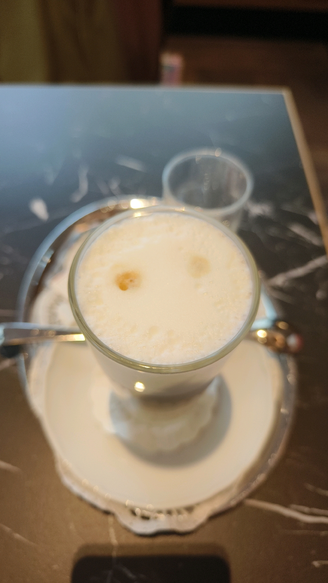 A close-up, overhead shot shows a glass of foamy milk, possibly a latte or cappuccino, placed on a white saucer. The drink has two small, light brown spots on its surface, resembling a smiley face. The glass and saucer are on a silver tray with intricate detailing. In the background, another empty glass is visible, and the surface appears to be a dark, marbled table. The scene is likely in Vienna, Austria, suggesting a setting within a cafe or restaurant. No people are visible in the image, and no specific actions or interactions are depicted.