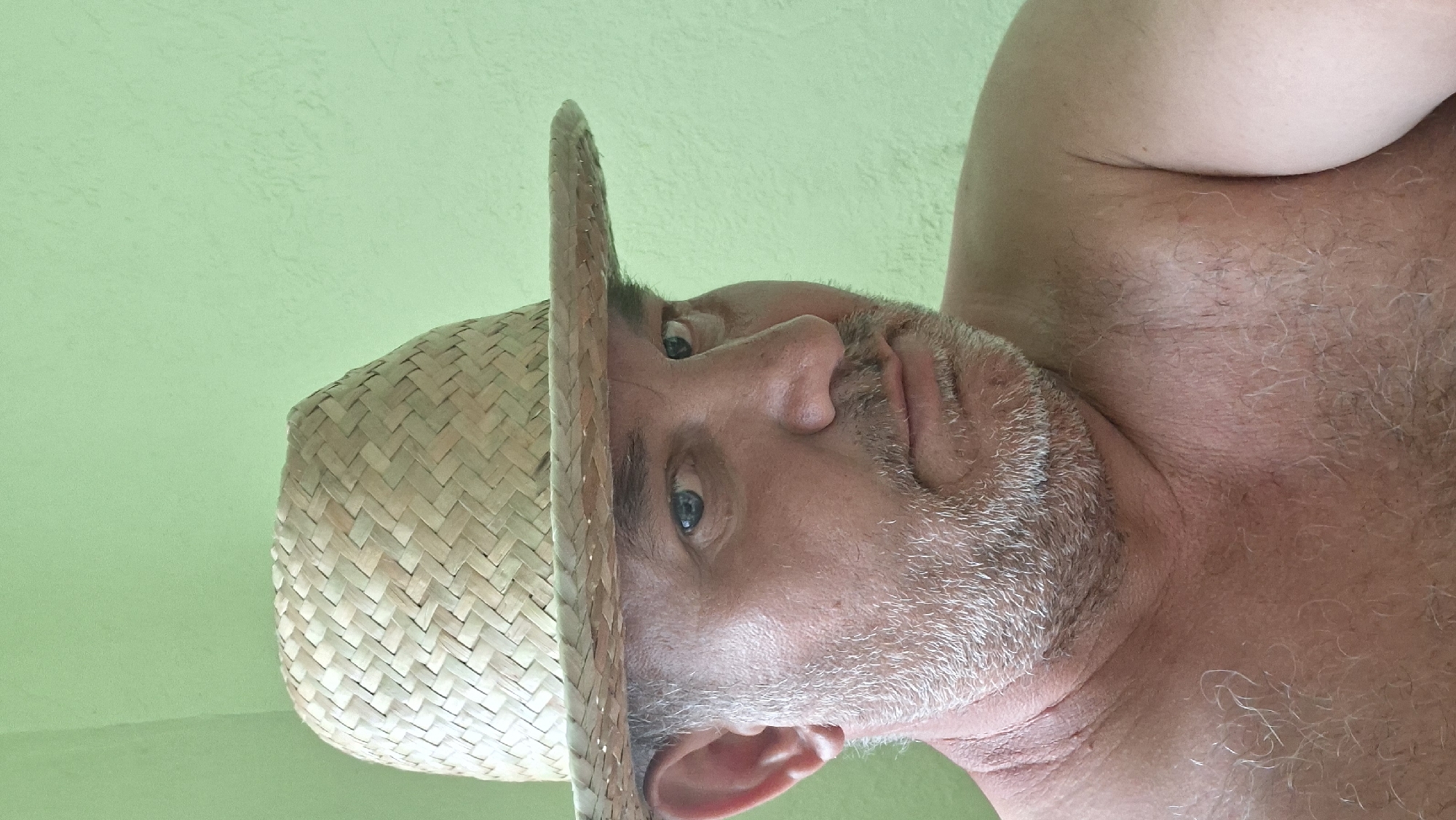 A close-up, chest-up portrait of a middle-aged man wearing a straw hat, taken indoors against a light green textured wall. The man has short, graying hair, a graying beard, and blue eyes. He is looking slightly off-camera with a neutral expression. His chest and shoulders are visible, and his skin appears slightly reddened. The hat is tilted forward, casting a shadow over the upper part of his face. The background is a plain, light green wall. There are no discernible elements to indicate the location as Voinova, Moldova.