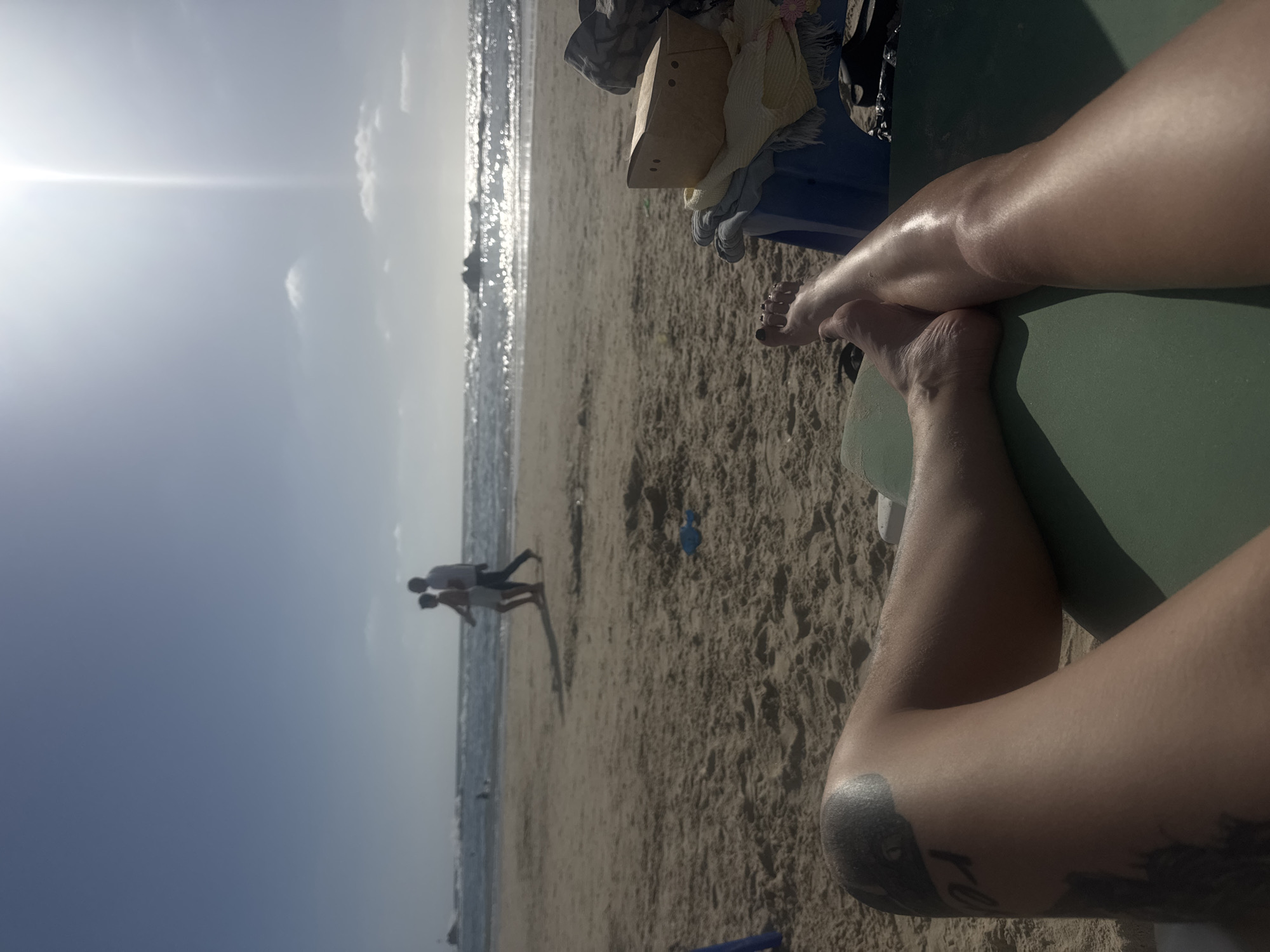 The image captures a beach scene in Bat Yam, Israel. A pair of legs, one bearing a tattoo, are resting on a green surface, likely a beach chair or towel. Personal belongings sit nearby. In the background, two individuals are walking on the beach towards the ocean, their figures silhouetted against the bright sunlight reflecting on the water. The sky is blue with a few clouds, suggesting a sunny day. The beach appears sandy, with a blue object (possibly a ball) visible in the foreground.