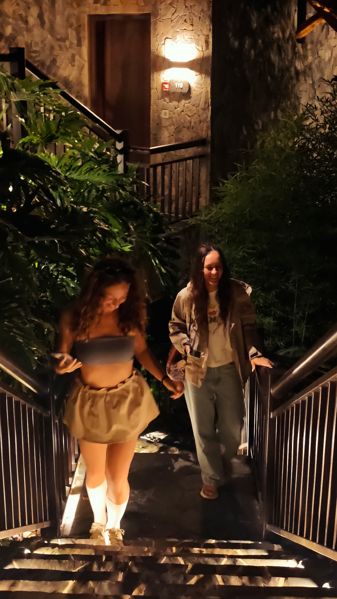 The image captures two females descending an outdoor staircase at night within a resort or homestay setting in Mu Cang Chai, Vietnam.
On the left, positioned lower on the stairs, is a female with long, curly dark brown hair. She is wearing a light blue strapless crop top, a light tan voluminous skirt, white knee-high socks, and light-colored lace-up shoes. She looks down and to her right with a gentle smile, her left hand near her torso and her right hand slightly extended.
Behind her, on the right side of the stairs, is a second female with long, dark, wavy hair. She is wearing a light-colored t-shirt with a visible graphic design, an open light brown or beige oversized jacket, light blue jeans, and light-colored open-toed sandals. She is smiling and looking slightly forward, with her right hand resting on the dark metal railing and her left hand holding a small, possibly patterned, bag or object close to her body.
The staircase consists of dark steps and features dark metal railings with vertical balusters on both sides. The surrounding structures are clad with irregular, brownish-grey stone blocks. Warm yellow light emanates from two wall-mounted sconces on the stone wall at the top of the stairs. Below the sconces, a wooden door is set into the wall, and a light-colored plaque clearly displays the room number "110". A small red rectangular object (likely a fire alarm pull station) and a white square electrical outlet or switch are also visible on the stone wall near the room number. Dense, dark green foliage, including broad-leafed plants on the left and thinner, possibly bamboo-like stalks on the right, flanks the staircase. The scene is illuminated by artificial lights, casting shadows and indicating nighttime conditions.