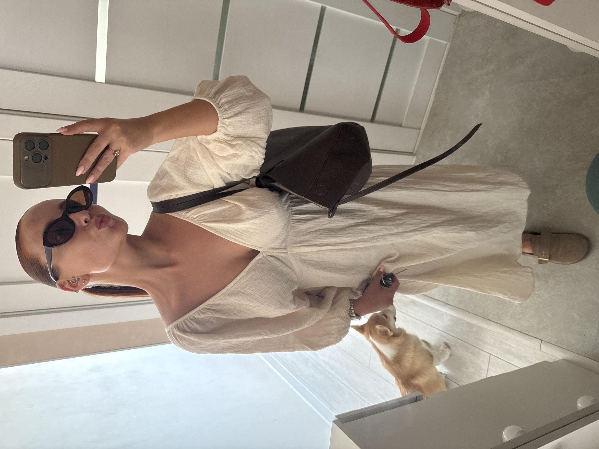 A woman is taking a selfie indoors. She is wearing a light-colored dress, sunglasses, and has a brown purse slung over her shoulder. She is holding a phone with a brown case. A small dog is near her feet. The background shows a white door and wall. The location is not discernible as the image does not contain features specific to the city of Chaikovsky, Russia.