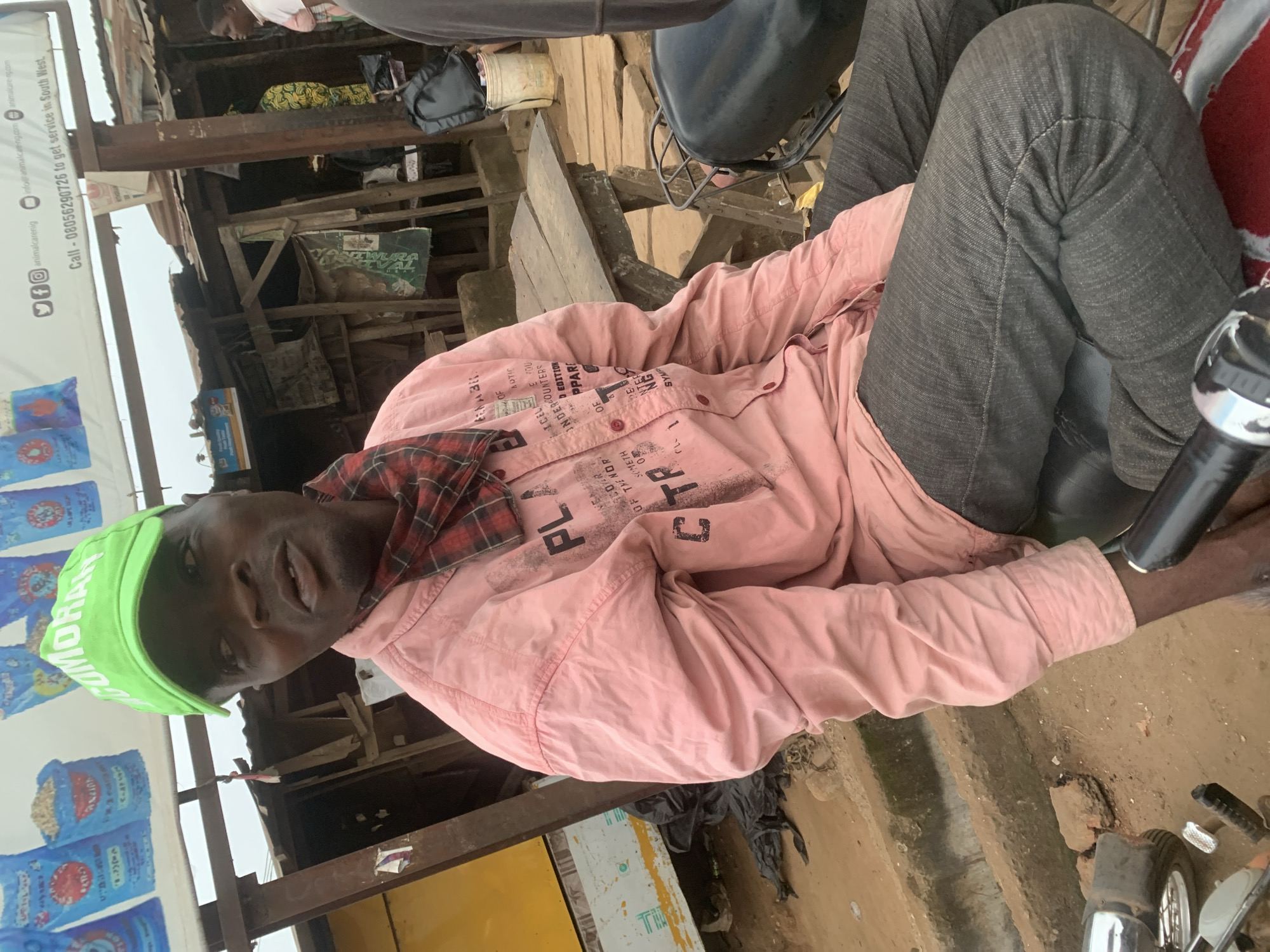 In Shagamu, Nigeria, a dark-skinned man wearing a bright green cap with white lettering, a pink long-sleeved shirt with black text, and dark grey jeans is seated on a motorcycle. He is looking to his right, with his mouth slightly open. A red and black checkered scarf is tied around his neck. The background shows a rustic outdoor market setting with wooden stalls and corrugated metal roofing. Several bags are visible hanging from a structure above, one of which has blue and white branding. A wooden table is set up in front of one of the stalls. The ground is dusty and uneven. The overall impression is a candid shot of an individual in a busy, informal commercial area.
