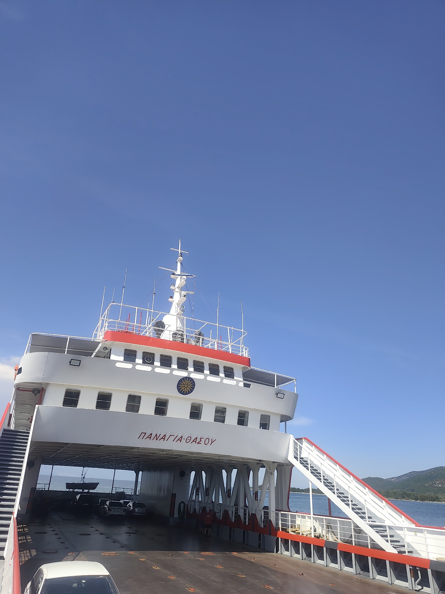 A low-angle, slightly tilted full shot shows the upper decks and the open car deck of a white ferry with a red stripe under a clear blue sky. The ferry is identified by red Greek lettering on its side, reading "ΠΑΝΑΓΙΑ-ΘΑΣΟΥ". Two sets of white stairs with red railings lead up the sides of the ferry. Several cars are visible on the open car deck. The water of a bay is visible through the opening of the car deck, with a distant green, tree-covered hillside. A small, dark rowboat is suspended from the ceiling of the car deck. The sun is visible as a yellow emblem with a blue background on the side of the ferry.