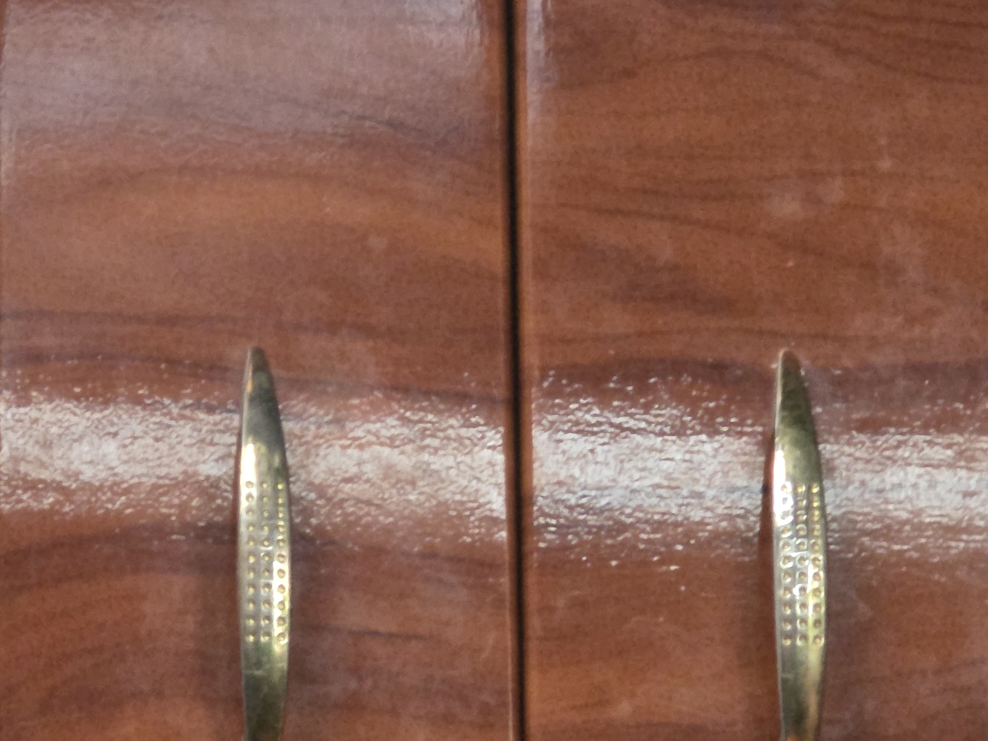 The image shows a close-up of a polished wooden surface with two brass drawer pulls. The drawer pulls are identical and are positioned horizontally. They are elongated with a slightly raised center that is decorated with a pattern of small dots. The wood grain is visible and has a reddish-brown hue, with a high gloss finish that reflects light. A thin horizontal line, likely a seam between drawers, is visible in the center of the image. The lighting is bright and creates glare on the surface. There are no people or other objects visible in the frame. The location is unspecified.